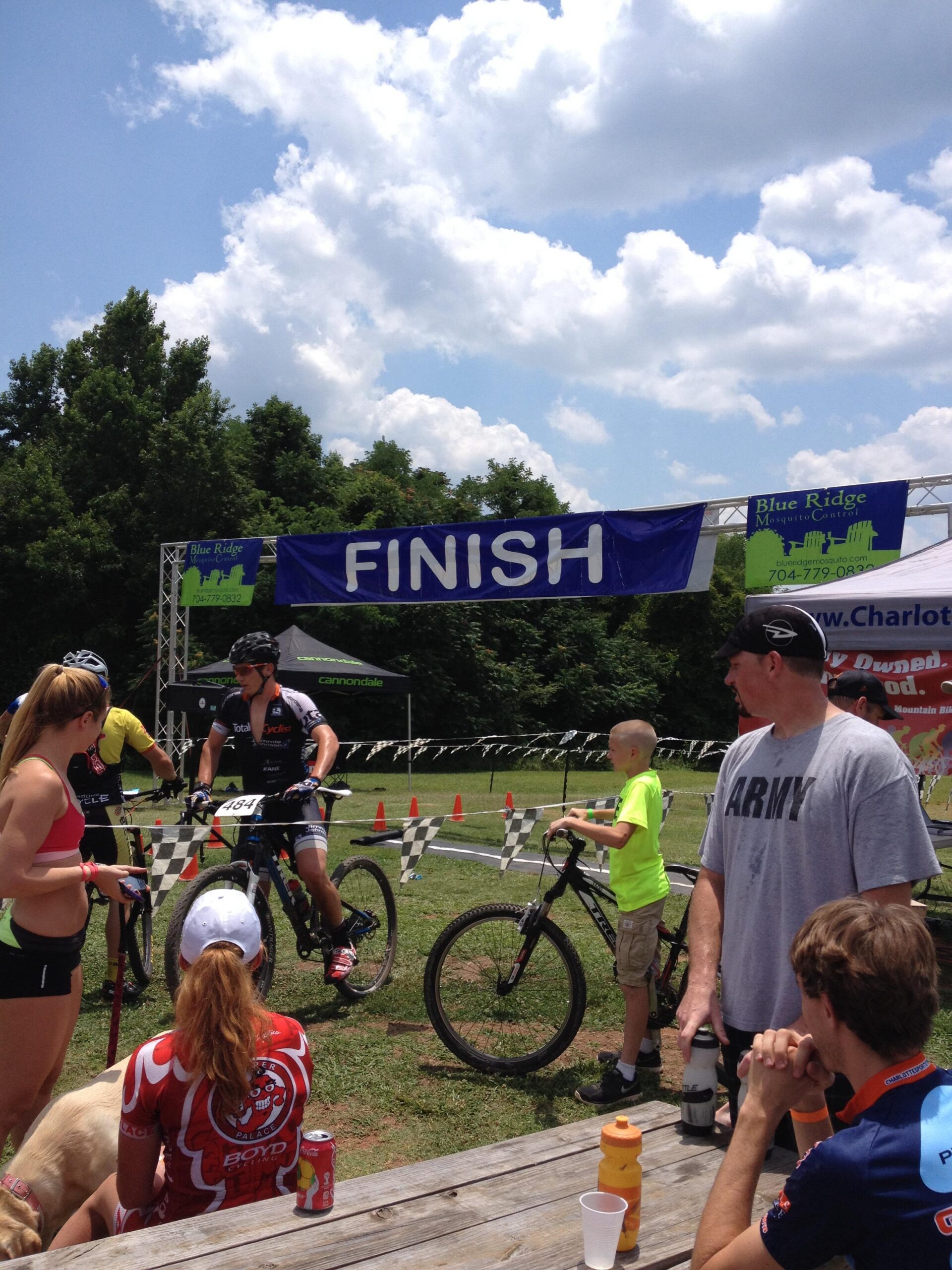 A mountain biking event finish line scene under a blue sky with fluffy clouds. A large "FINISH" banner is displayed, while participants and spectators gather around. In the foreground, a diverse group of people relaxes at a wooden table, with some looking at racers crossing the finish line. A young boy in a green shirt is seen with a bicycle, and a person in a red cycling jersey sits with a dog nearby. Anne Springs Close Greenway mountain bike trail.