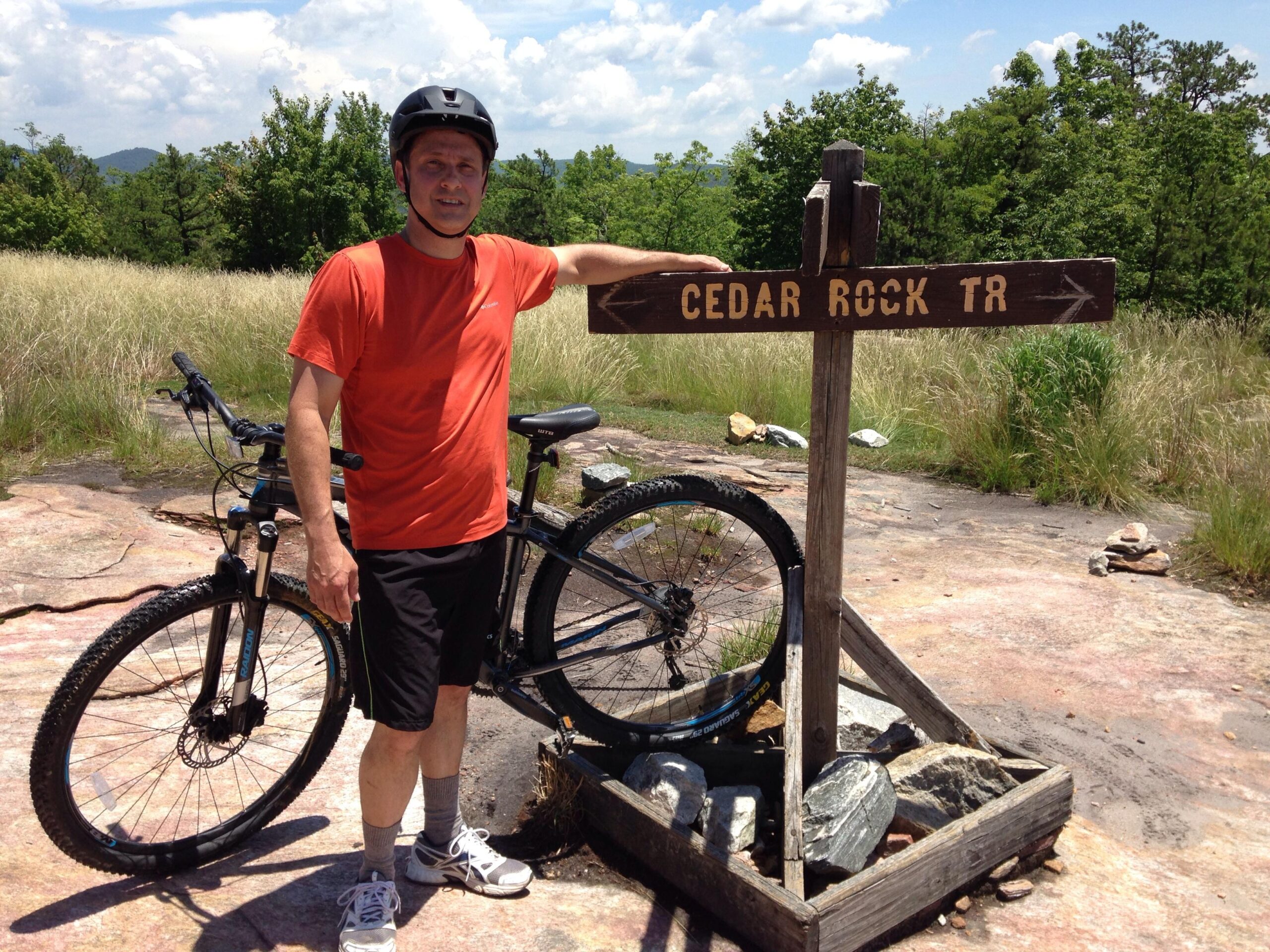 A person in an orange shirt and black shorts stands next to a wooden trail sign that reads "CEDAR ROCK TR." He leans against a mountain bike, with a grassy and rocky landscape in the background under a partly cloudy sky. Cedar Rock Trail #16 mountain bike trail.