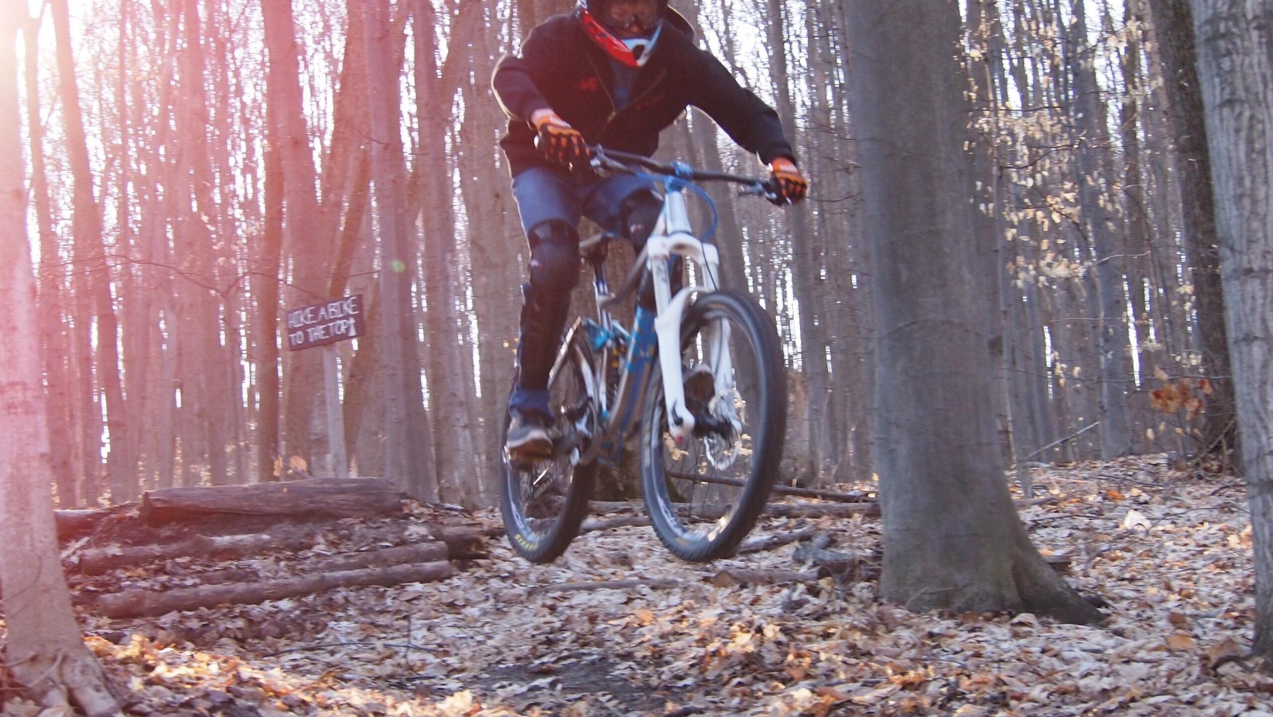 A mountain biker in protective gear jumps over a log on a wooded trail, with a sign in the background indicating the route to the top. The forest is filled with bare trees and fallen leaves, and the scene is illuminated by soft sunlight. Copper Ridge Freeride mountain bike trail.
