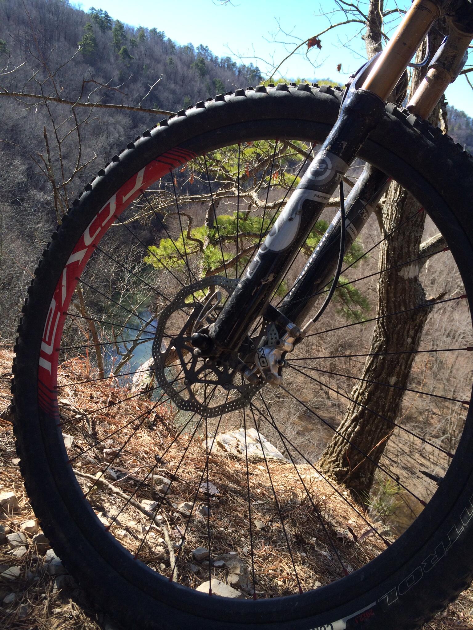 Specialized Camber Expert Carbon 29: A close-up view of a mountain bike wheel positioned at the edge of a cliff, overlooking a river below. The surrounding landscape features trees and rocky terrain, with a clear blue sky in the background.