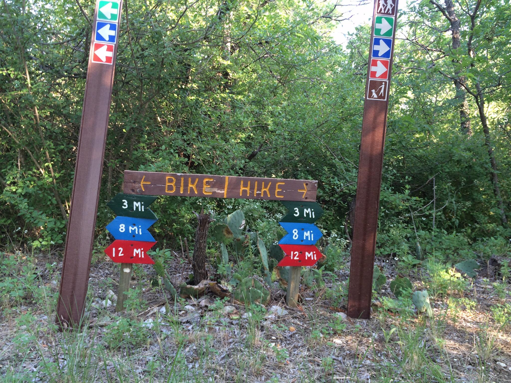 A wooden trail sign indicating directions for biking and hiking, with arrows pointing left and right, and distances marked in miles (3, 8, and 12) on colored signs below. Surrounded by green foliage in a natural setting. Cedar Hill State Park At Joe Pool Lake mountain bike trail.