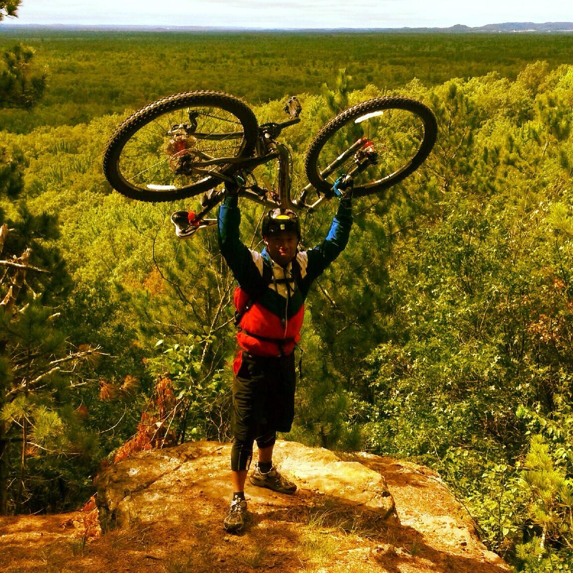 A person wearing a helmet and a colorful jacket stands on a rocky outcrop, triumphantly holding a mountain bike above their head. The background features a lush, green landscape stretching into the distance under a partly cloudy sky. Levis Mounds mountain bike trail.