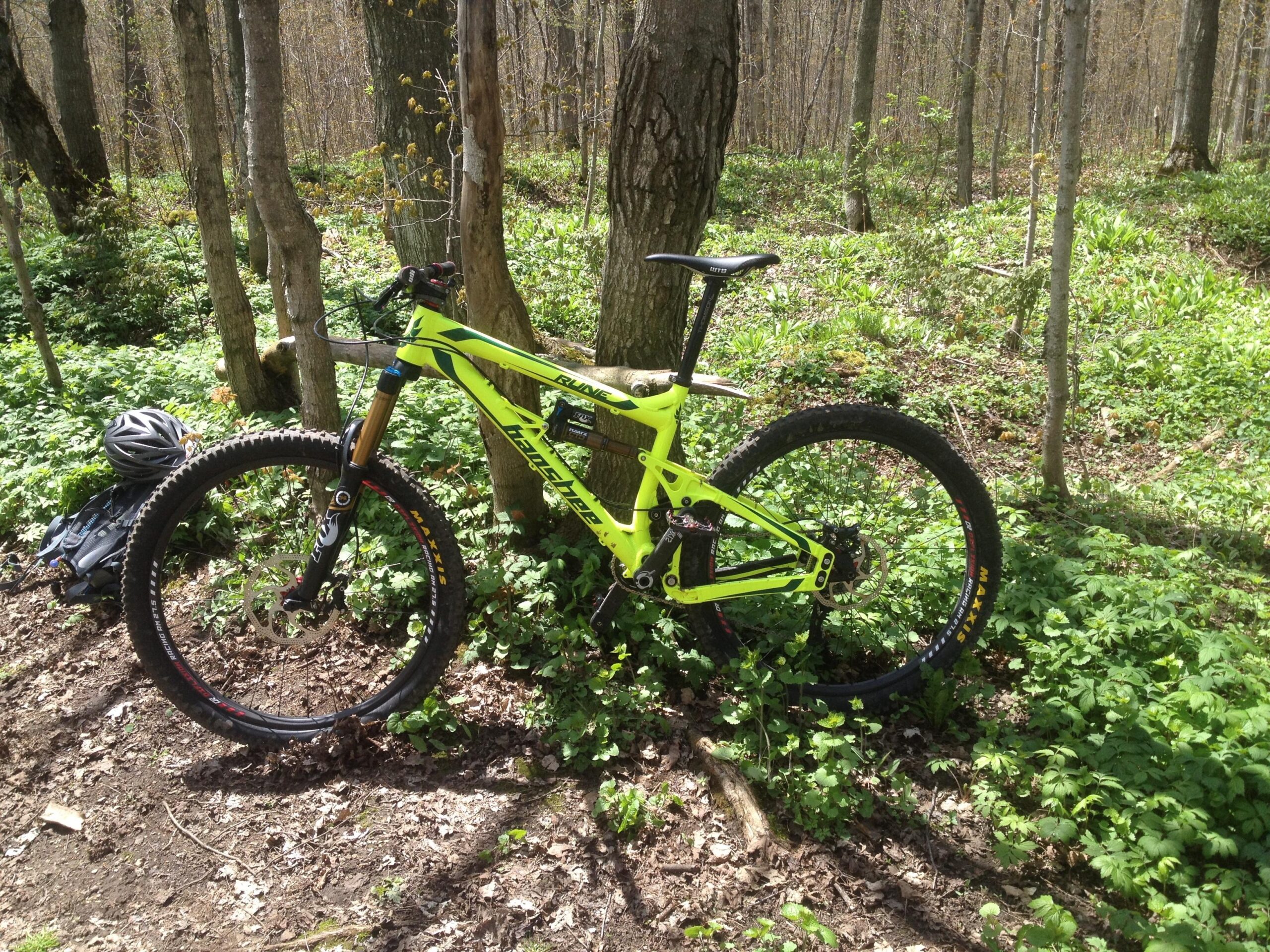 Banshee Rune: A bright yellow mountain bike leaning against a tree in a forested area, surrounded by lush green foliage. Nearby, a black helmet and backpack are placed on the ground.