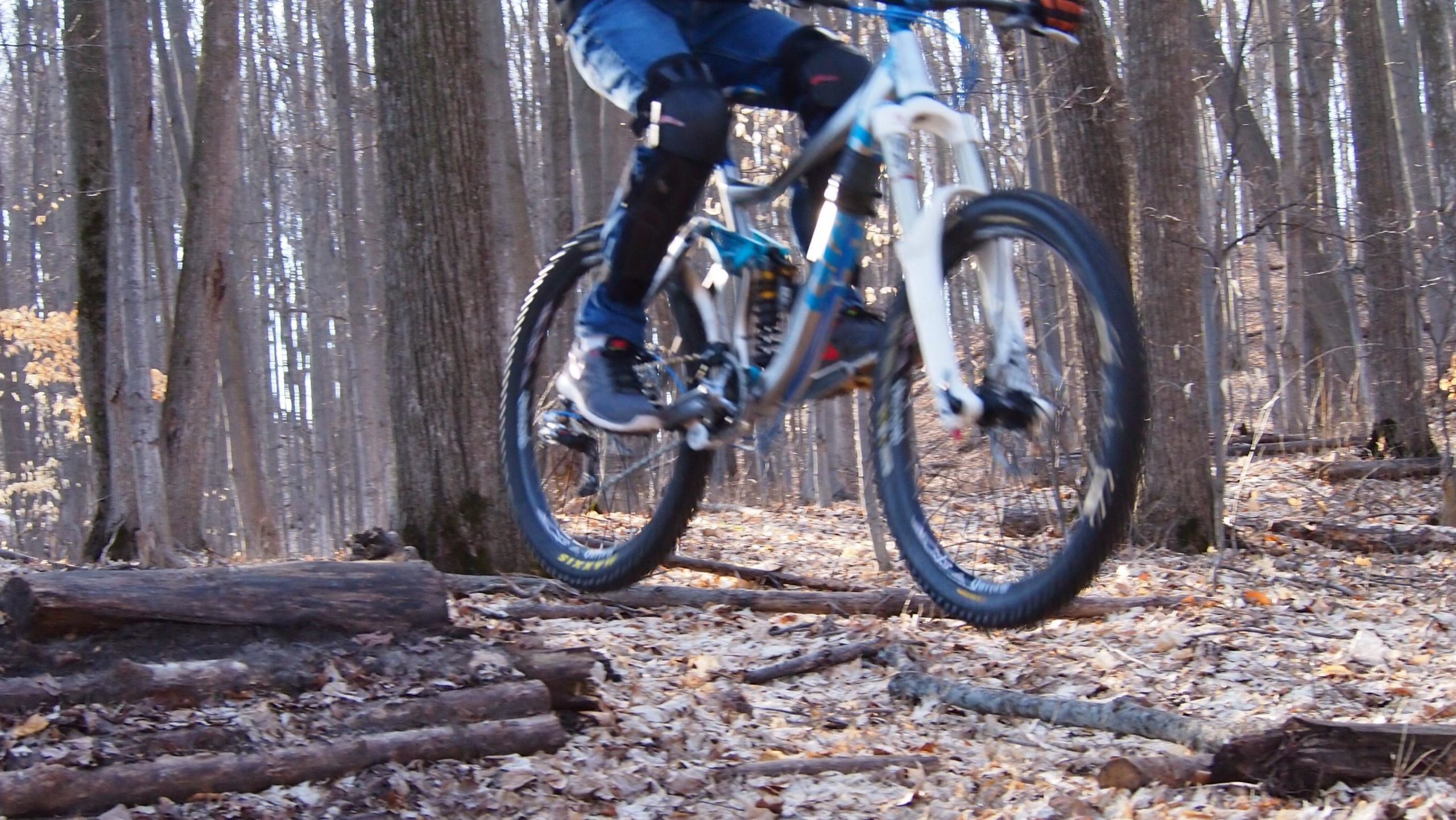 A mountain biker in protective gear jumps over a log on a forest trail covered with fallen leaves, surrounded by tall trees in the background. Copper Ridge Freeride mountain bike trail.