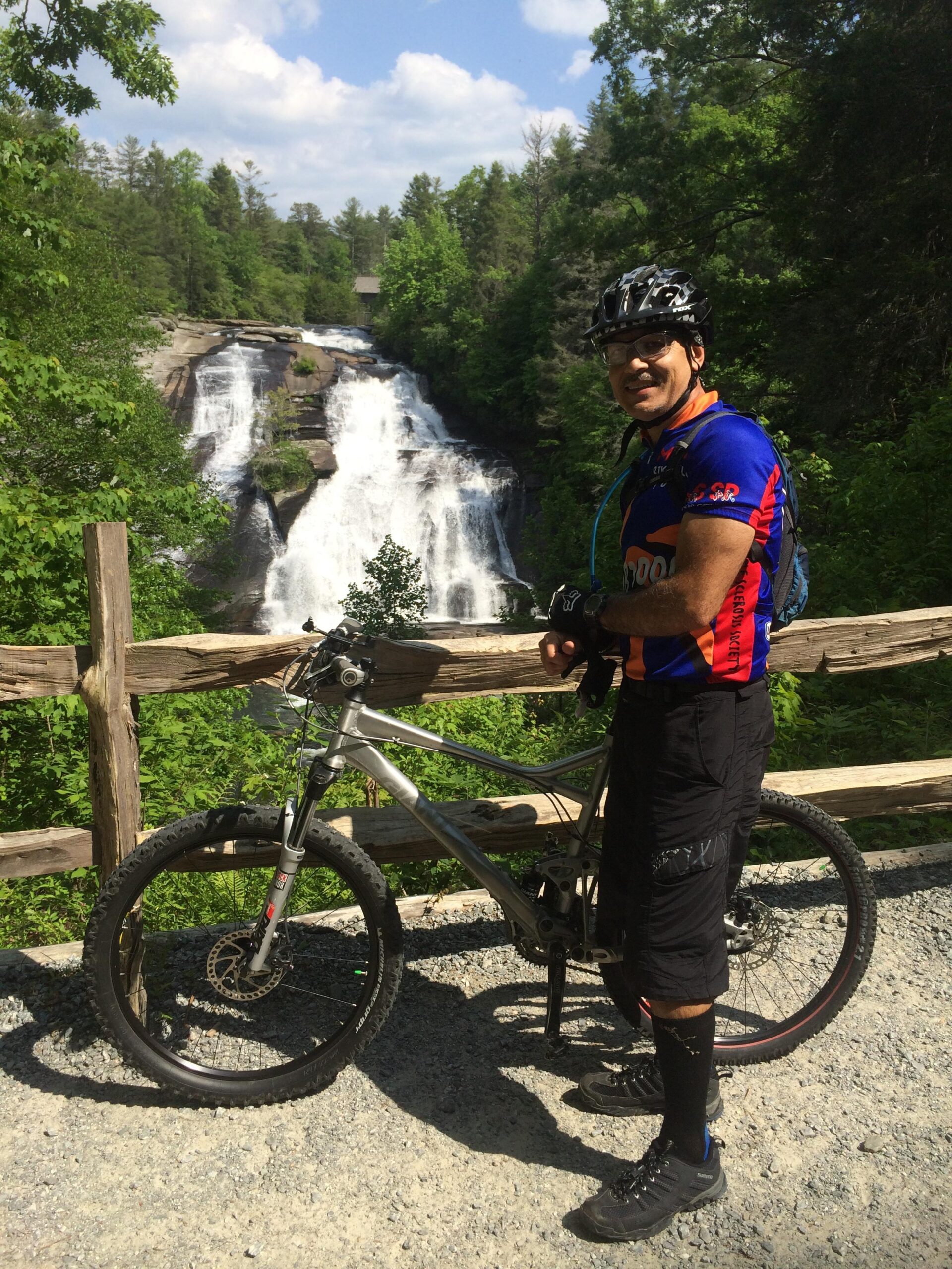 A cyclist stands next to a mountain bike on a gravel path, with a beautiful waterfall and lush greenery in the background under a partly cloudy sky. The cyclist is wearing a blue and red jersey, black shorts, and black socks, smiling at the camera. DuPont State Forest mountain bike trail.