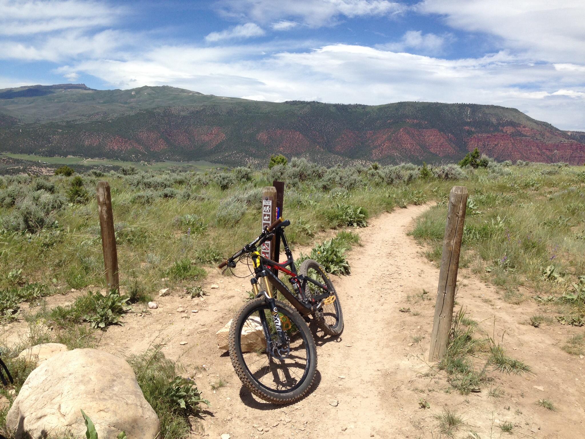 Devinci Dixon RS: A scenic view of a mountain biking trail with two parked bicycles in the foreground. The path is surrounded by green grass and shrubs, leading toward a mountainous landscape with red rock formations in the background under a partly cloudy sky. Trail markers are visible beside the bikes.