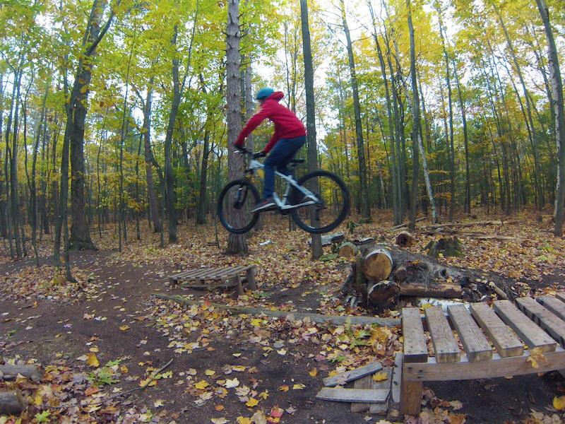 A person in a red jacket and blue helmet jumps a mountain bike over wooden obstacles in a forested area with autumn foliage. Copper Ridge Freeride mountain bike trail.