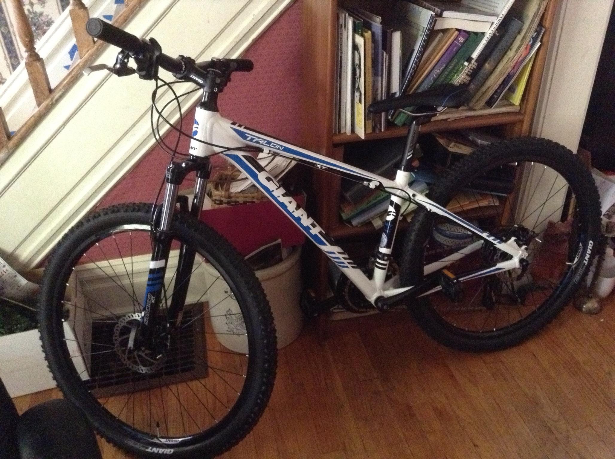 Giant Talon 27.5: A mountain bike leaning against a wooden bookcase, featuring a white frame with blue and black accents. The bike has thick tires and a front suspension fork, set on a wooden floor with various books and items visible in the background.