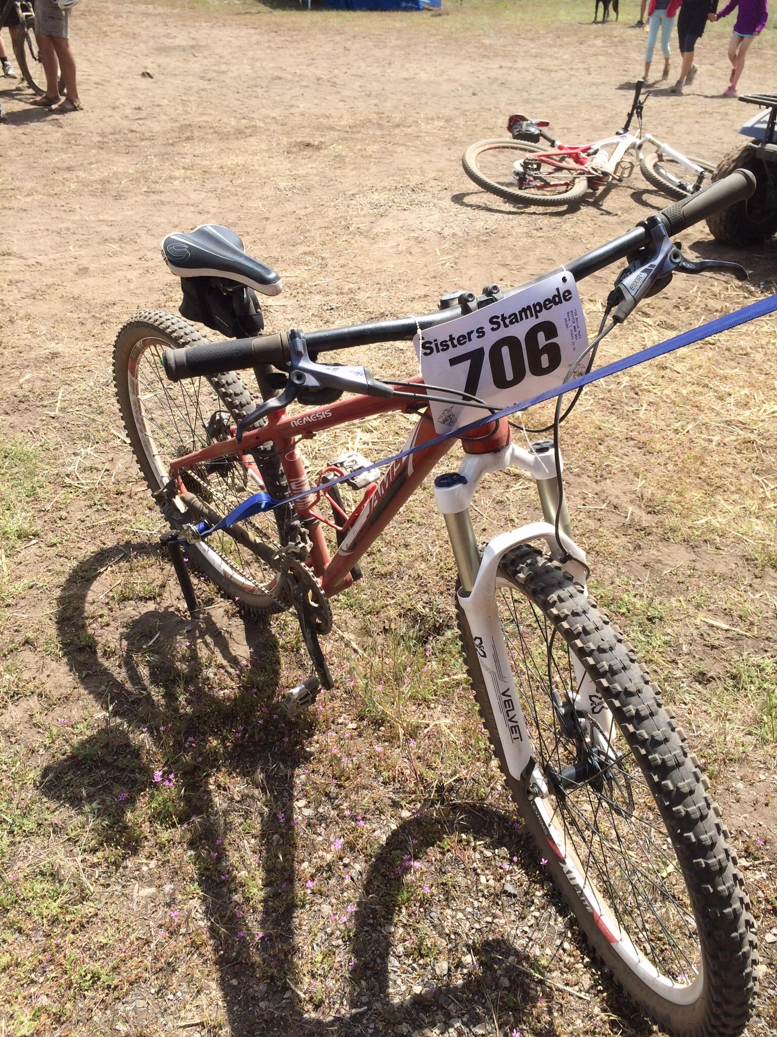 Jamis Nemesis: A mountain bike with a brown frame and a "Sisters Stampede" race number 706 attached to the handlebars. The bike is parked on a dirt surface scattered with small purple flowers, with other bicycles and people in the background.