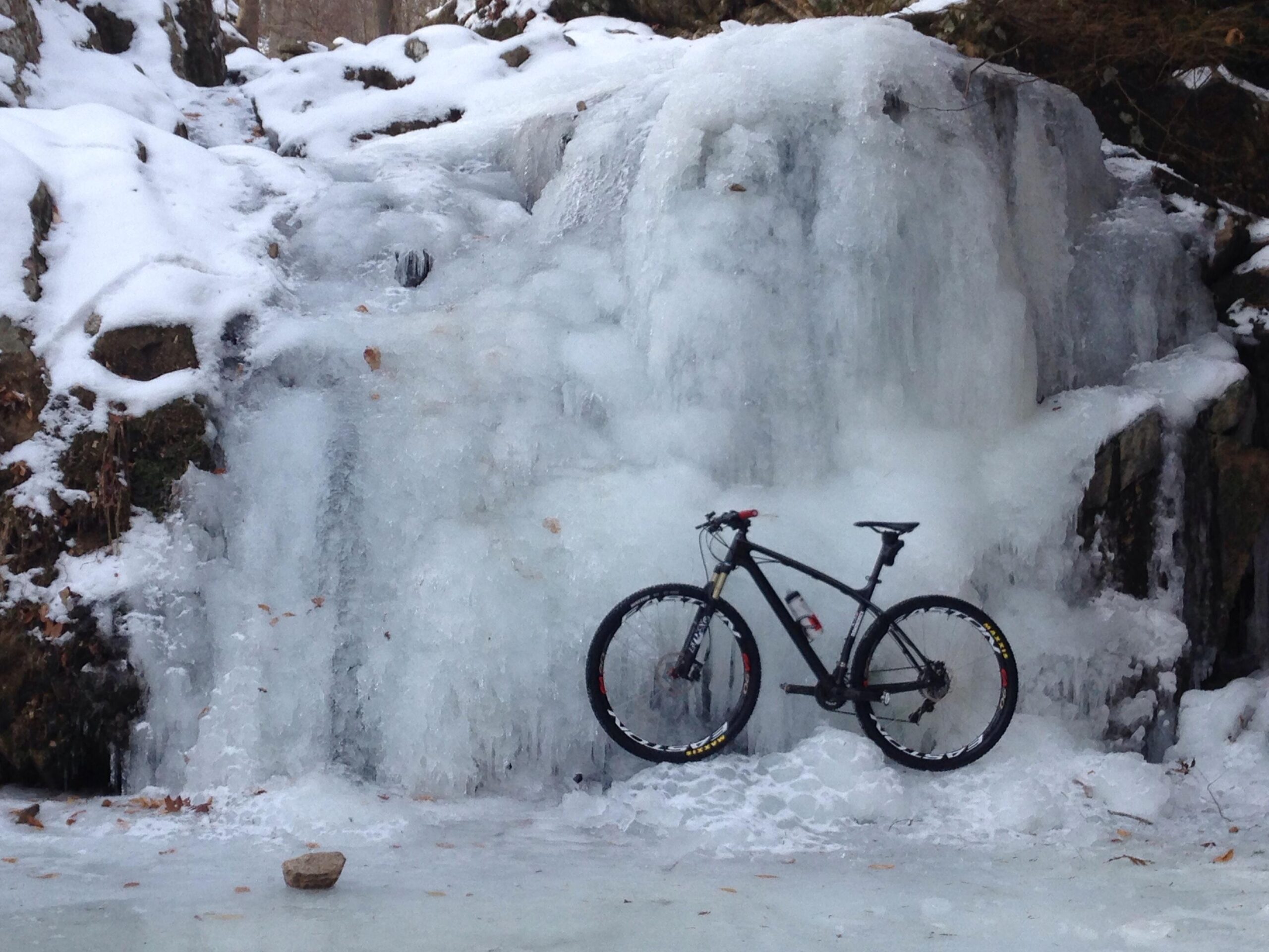 A mountain bike leaning against a frozen waterfall in a snowy forest setting. The scene features layers of ice and snow, with a small rock in the foreground. Patapsco Valley State Park (Avalon Area) mountain bike trail.