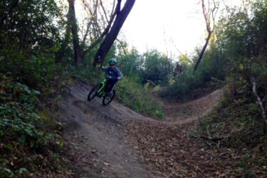 A child riding a green bicycle down a dirt trail in a forested area, surrounded by trees and autumn leaves. The child is wearing a helmet and appears to be navigating a small incline on the trail. Elm Creek Park mountain bike trail.