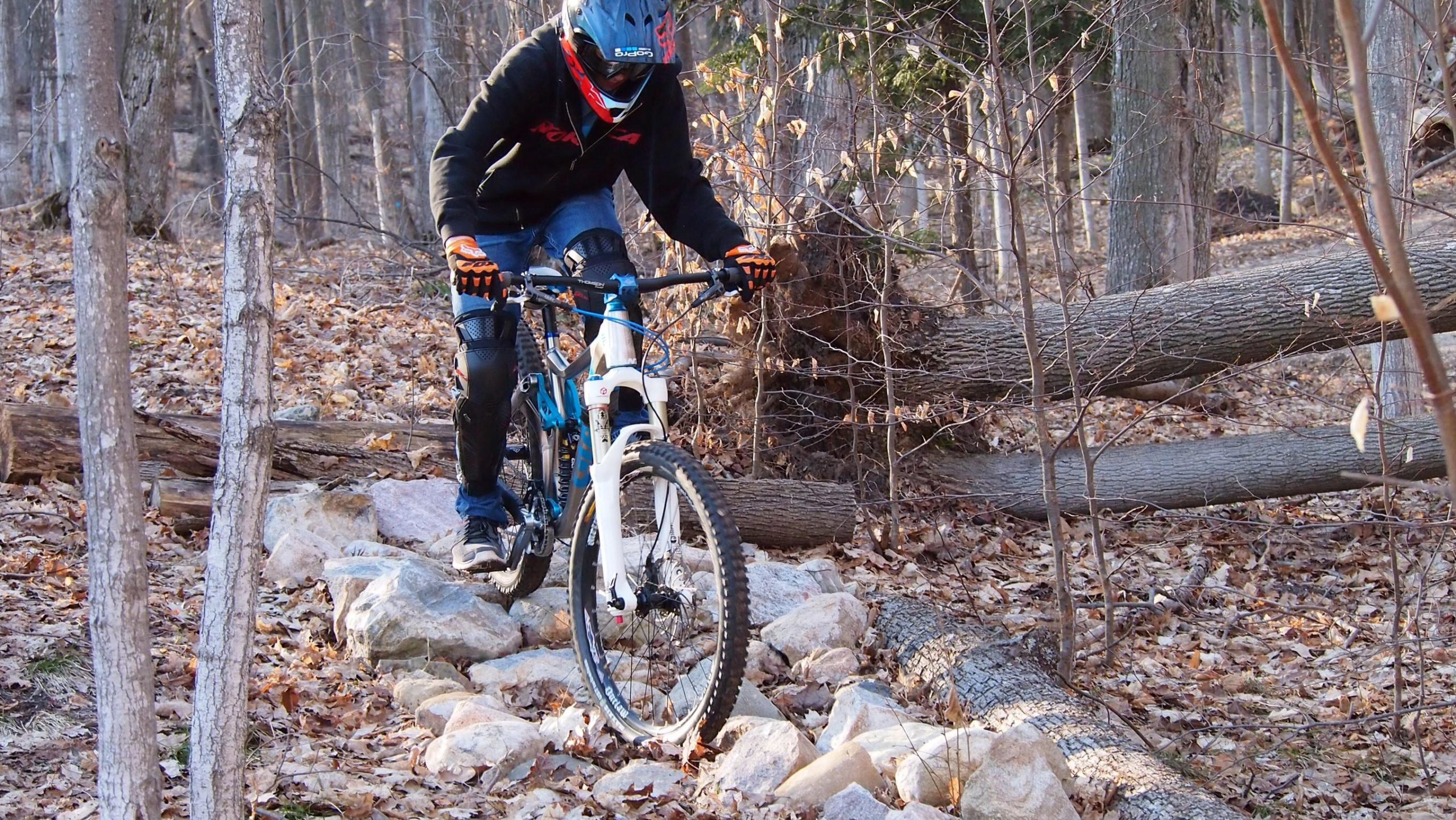 A mountain biker navigates rocky terrain in a forest during the fall, wearing a helmet and protective gear. The ground is covered with leaves, and there are fallen logs and rocks scattered throughout the scene. Copper Ridge Freeride mountain bike trail.