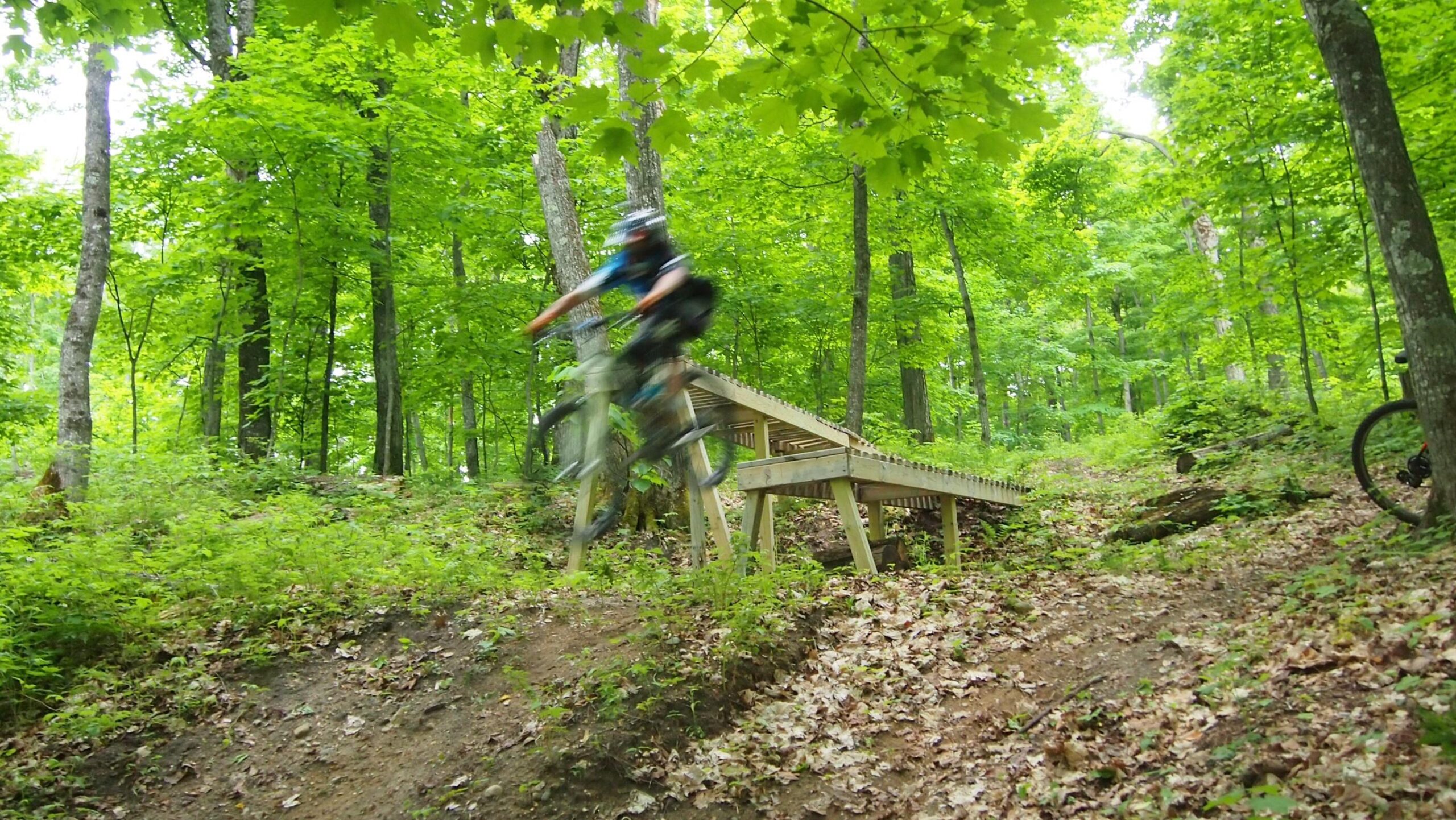 A mountain biker jumps off a wooden ramp in a lush green forest, surrounded by trees and foliage, with motion blur capturing the dynamism of the action. Copper Ridge Freeride mountain bike trail.