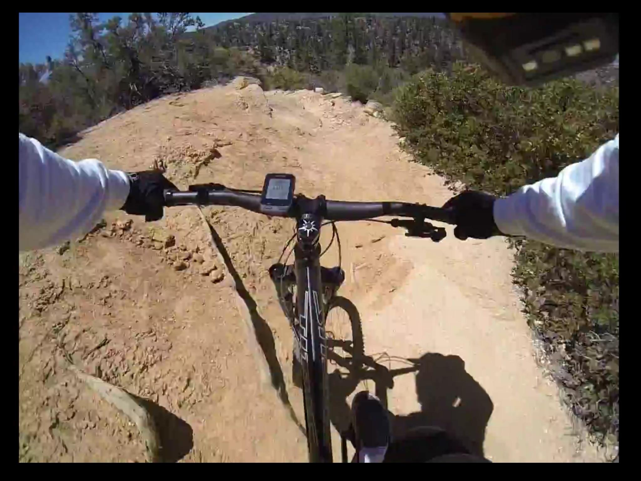 A view from above the handlebars of a mountain bike navigating a dirt trail surrounded by shrubs and trees. The cyclist is wearing gloves and a long-sleeve shirt, with a cycling computer mounted on the handlebars displaying information. The terrain is rocky and uneven, suggesting a challenging ride in a natural outdoor setting. Chilao / Silver Moccasin mountain bike trail.
