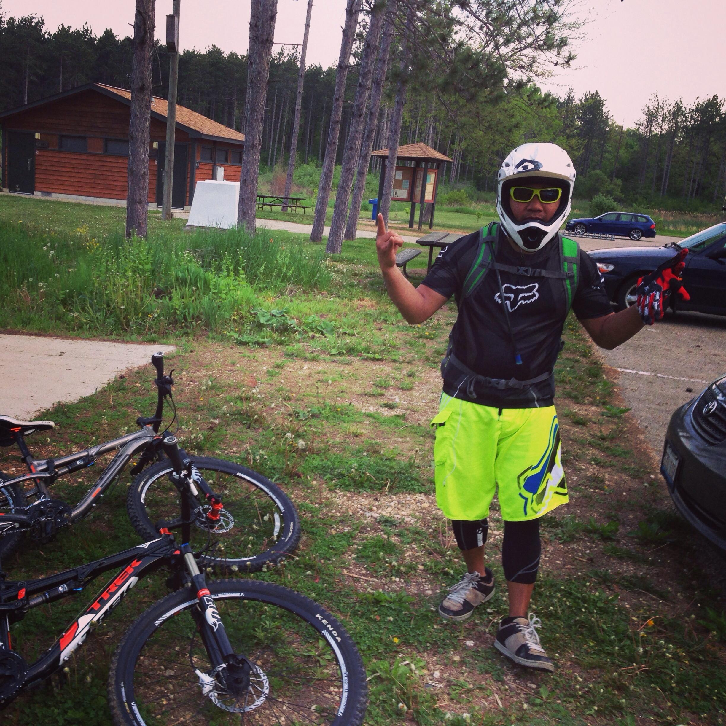 A person in a bright neon outfit and helmet stands next to two mountain bikes in a park setting. They are making a hand gesture while smiling, surrounded by trees and a wooden building in the background. Levis Mounds mountain bike trail.