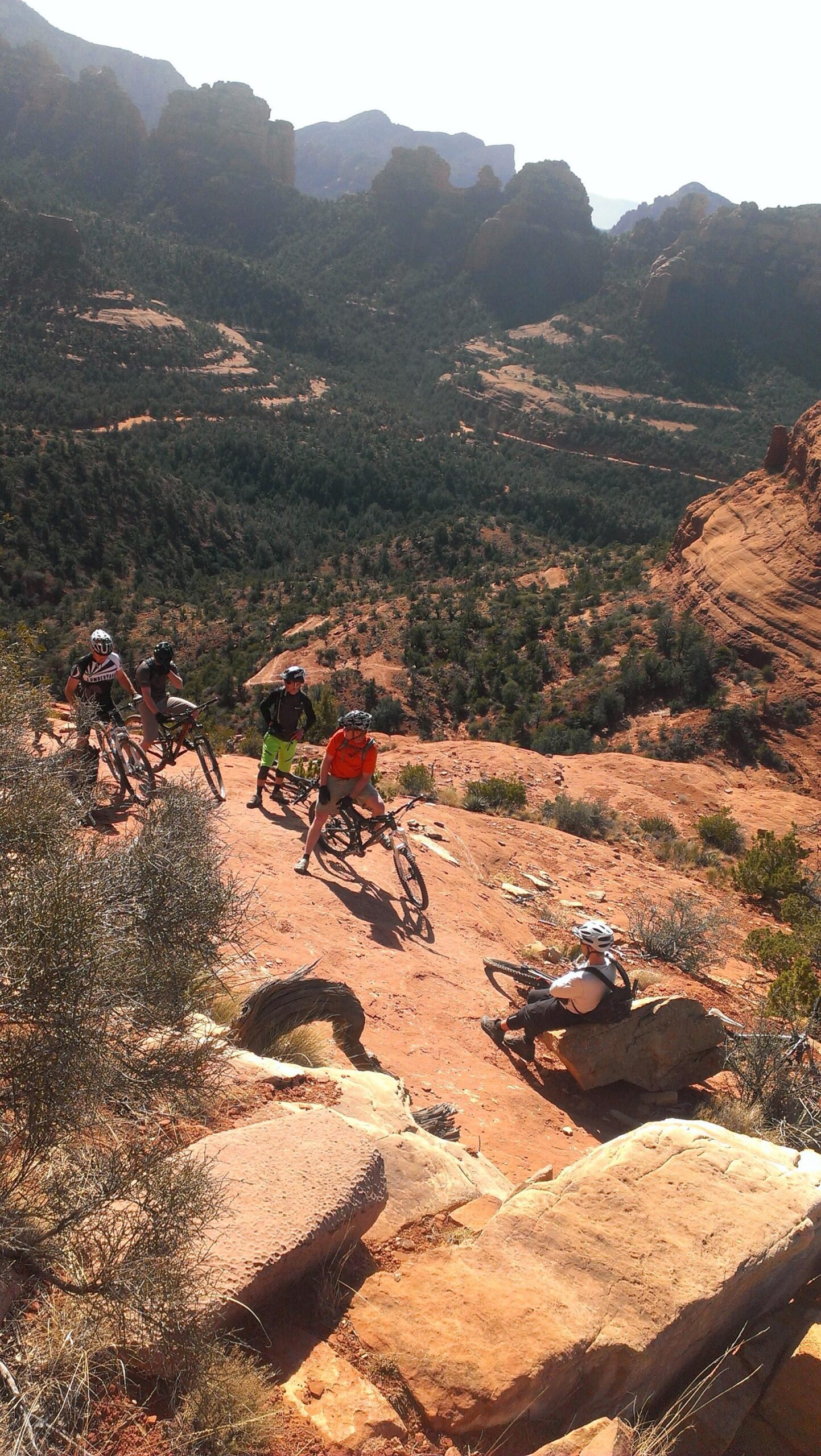 A group of mountain bikers navigating a rocky terrain in a desert landscape, surrounded by red rock formations and green vegetation. The backdrop features distant mountains and a clear sky. Hiline mountain bike trail.