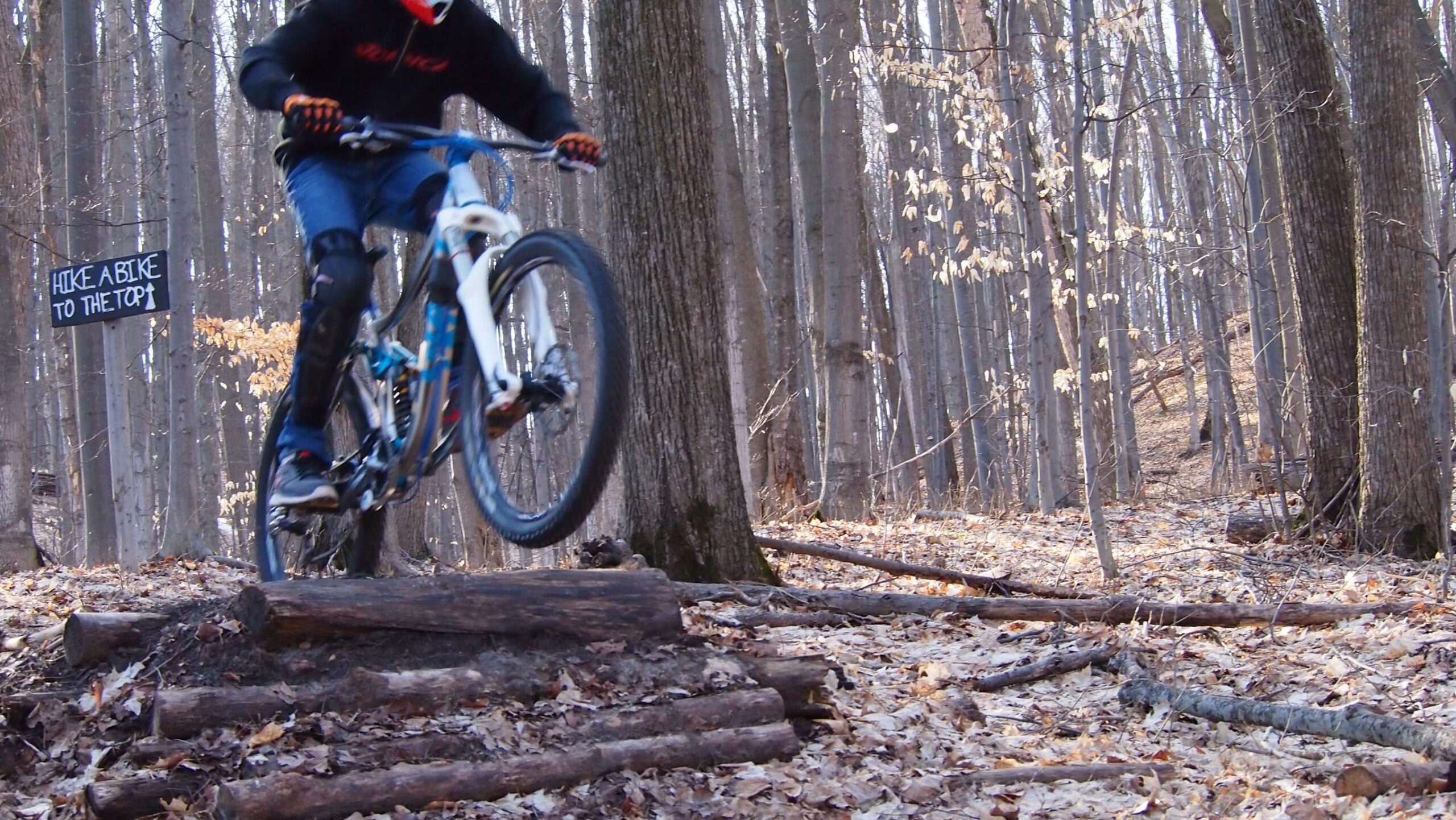 A mountain biker performing a jump over a series of wooden logs on a forest trail. The scene features trees in the background with autumn leaves scattered on the ground. A sign in the background reads "HIKE A BIKE TO THE TOP." The biker is wearing protective gear and a helmet. Copper Ridge Freeride mountain bike trail.