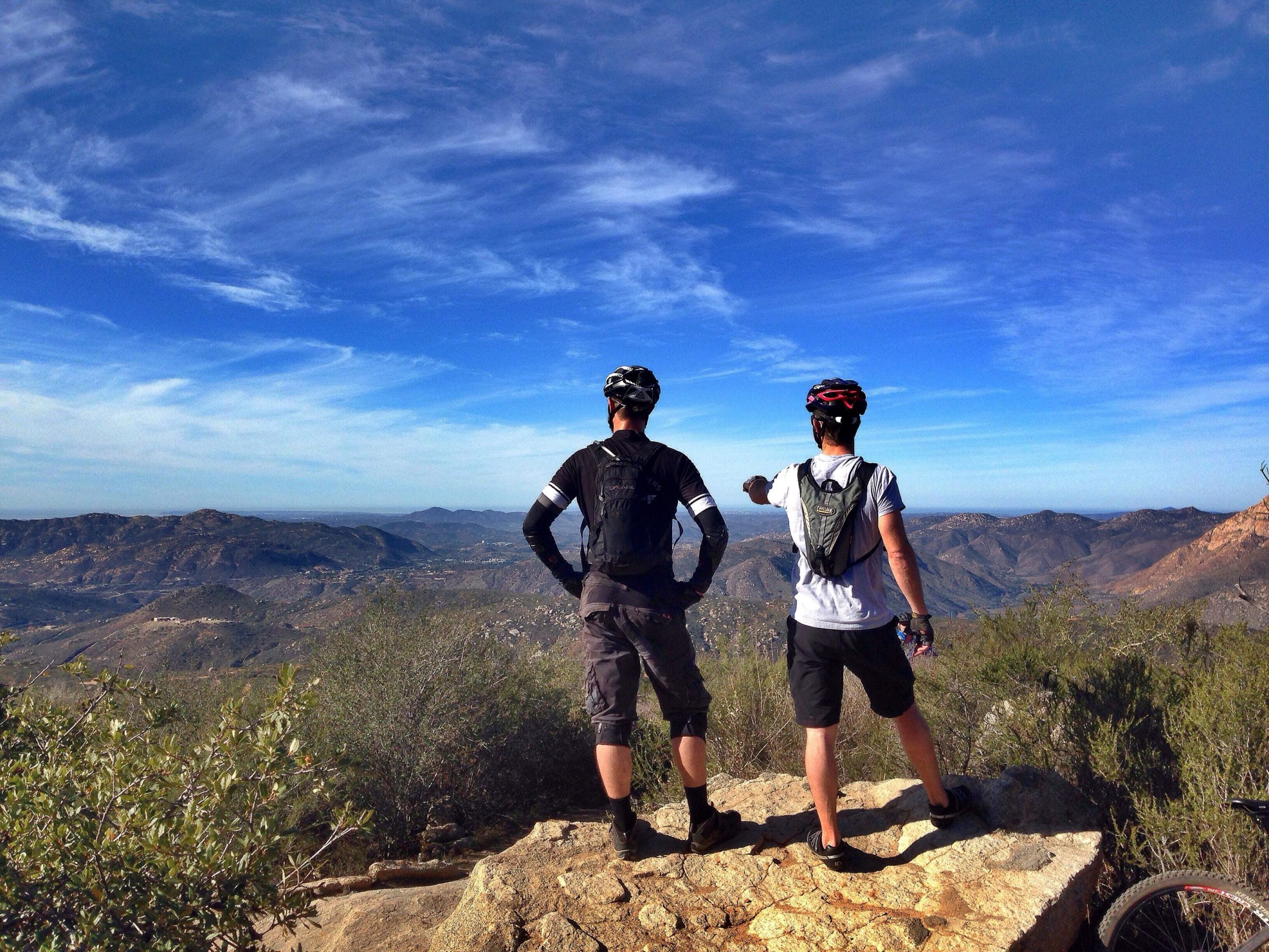 Two mountain bikers, standing on a rocky outcrop, admire a scenic view of rolling hills and valleys under a bright blue sky. One biker points out something in the distance while the other looks on, both wearing helmets and cycling gear. Lush greenery and shrubs surround them. Anderson Truck Trail mountain bike trail.