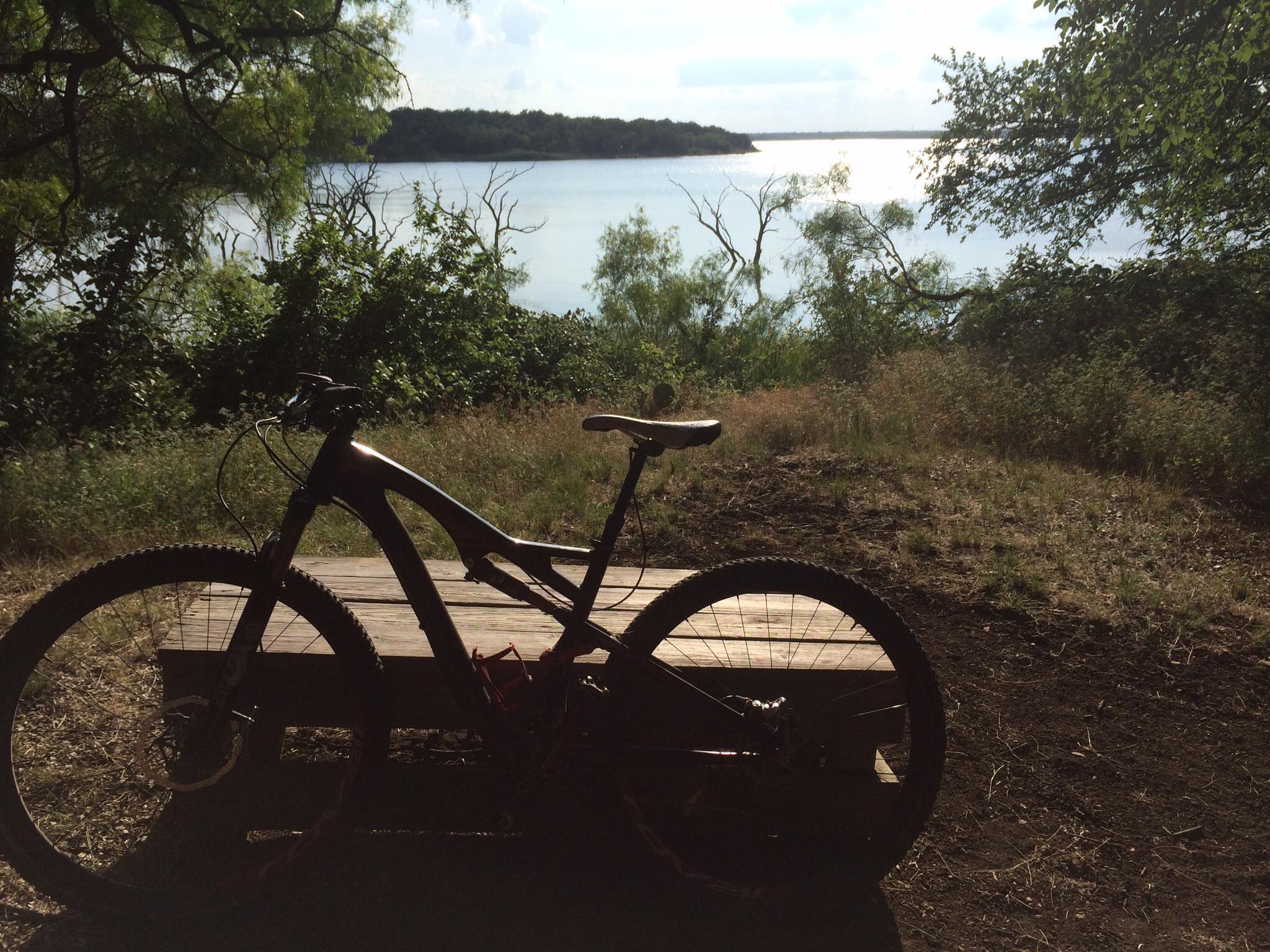 A mountain bike parked on a wooden platform overlooking a peaceful lake, surrounded by lush greenery and trees. The water reflects sunlight, creating a serene outdoor atmosphere. Cedar Hill State Park At Joe Pool Lake mountain bike trail.