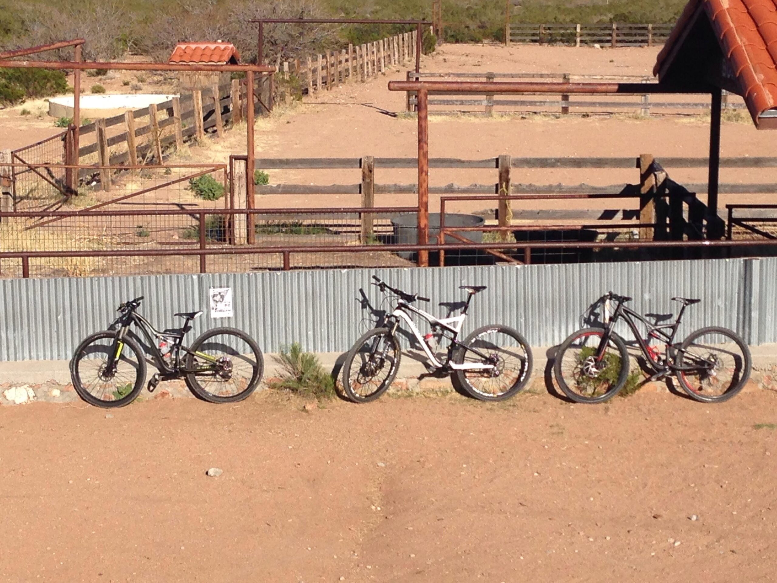 Specialized Camber Comp 29: Three mountain bikes parked next to a gravel path, with a rustic wooden fence and a partially covered area in the background. The setting appears to be an outdoor space, possibly near a ranch or recreational area, with sparse vegetation and a clear blue sky.