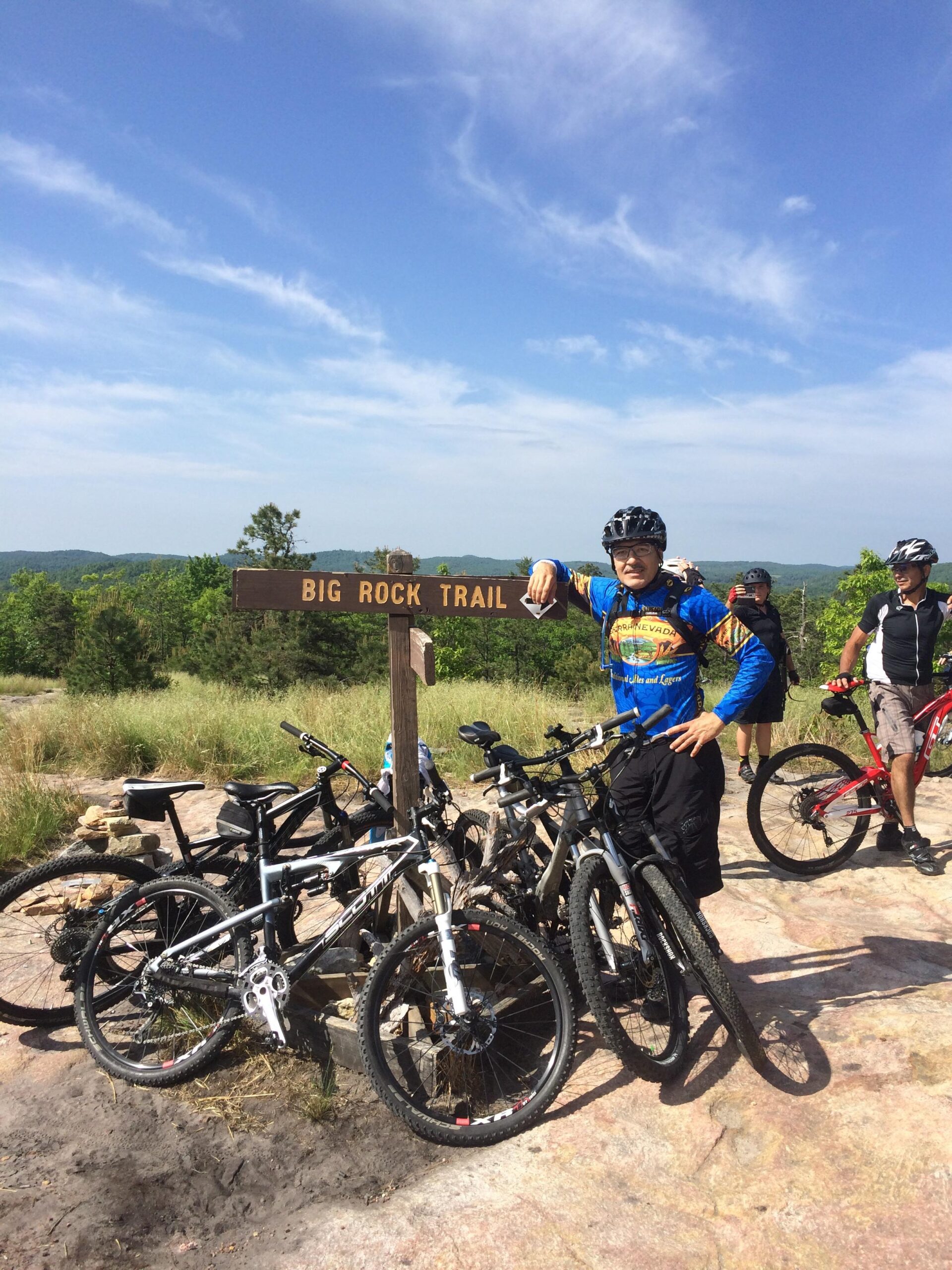 A group of mountain bikers resting at the Big Rock Trail sign on a sunny day. Several bicycles are parked nearby, and the landscape features green hills and a blue sky with a few clouds. One cyclist in a colorful jersey is posing with a smile while others enjoy the scenery in the background. DuPont State Recreational Forest mountain bike trail.
