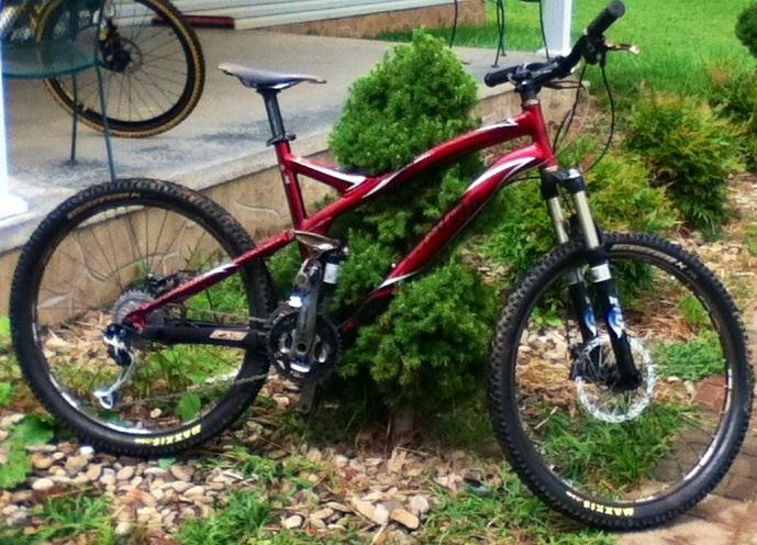 A red mountain bike is parked next to a small bush in a grassy area, with a porch visible in the background. The bike features sturdy tires, a dual suspension frame, and a modern design, indicating it's built for off-road riding. Flow mountain bike trail.