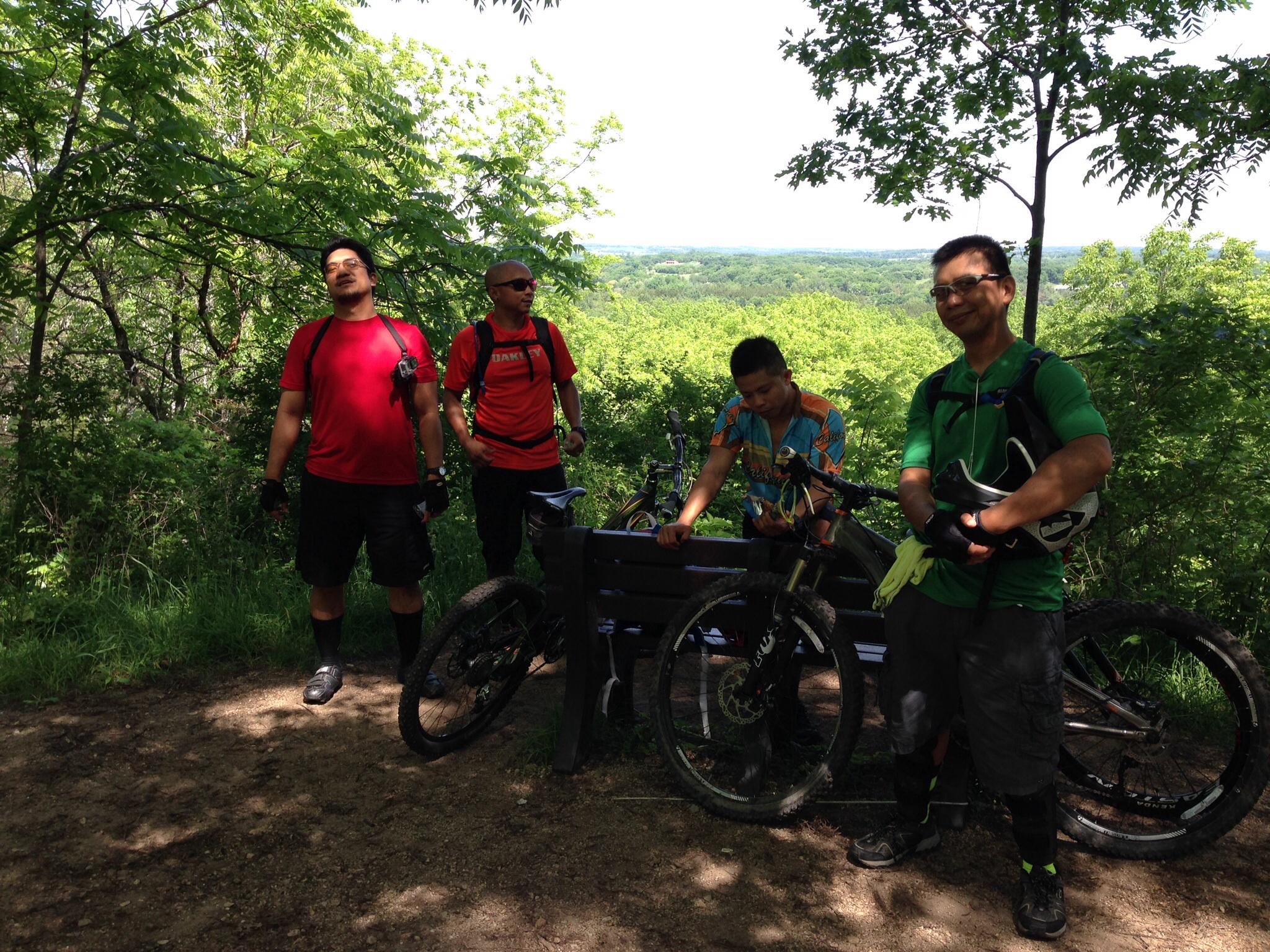 A group of four mountain bikers taking a break at a scenic overlook, surrounded by lush greenery. Two riders are standing with their bikes, while a third is seated on a bench and one is looking around. They are dressed in casual athletic gear, with one wearing a vibrant patterned shirt. The landscape behind them features rolling hills and trees under a clear sky. Emma Carlin mountain bike trail.