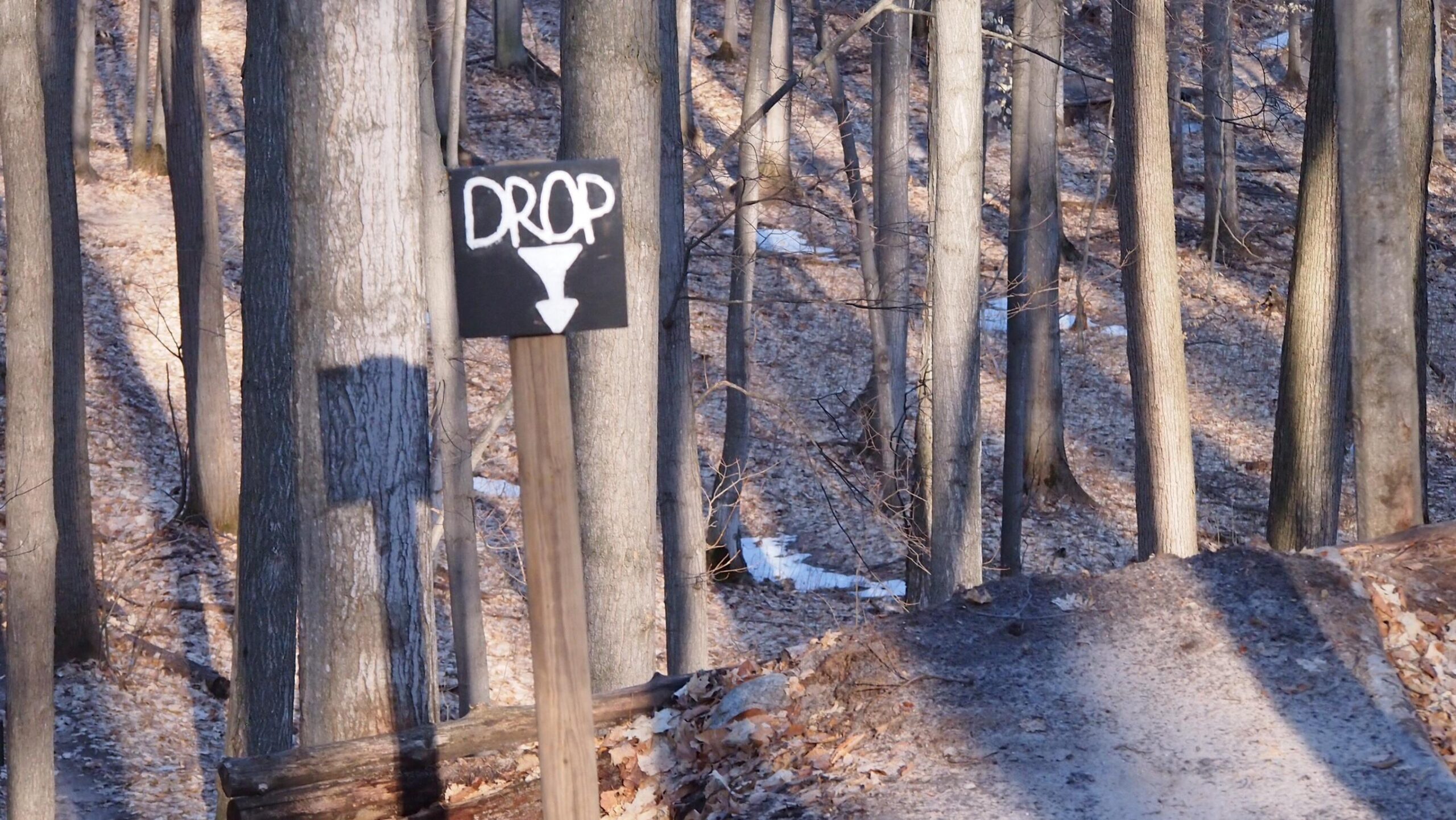 A wooden sign labeled "DROP" with an arrow pointing downwards is positioned on a dirt path through a wooded area. Tall trees surround the scene, with fallen leaves covering the ground and remnants of snow visible in the background. The sunlight creates a play of shadows among the trees. Copper Ridge Freeride mountain bike trail.