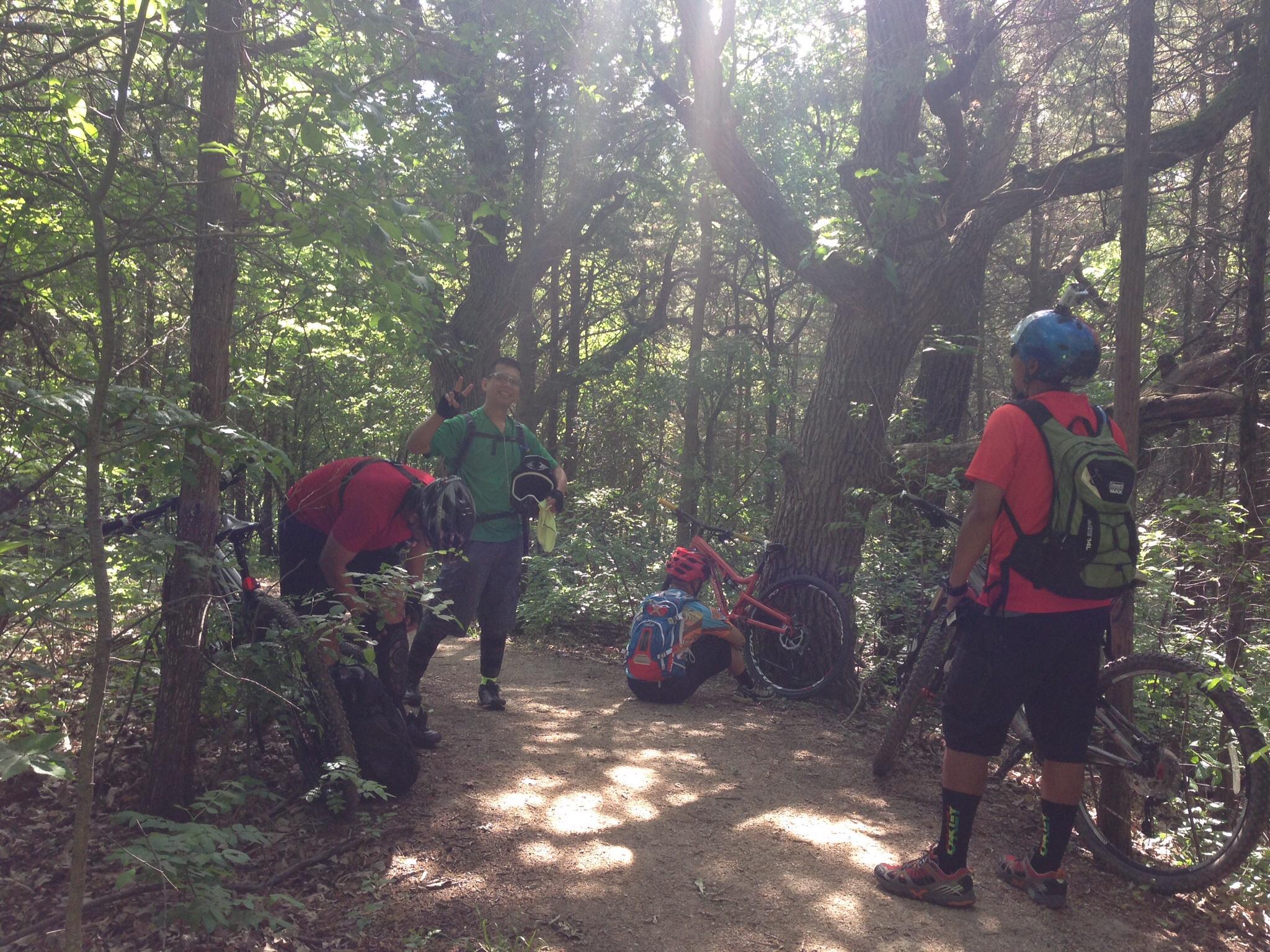 Four mountain bikers taking a break in a wooded area. Two are standing near their bikes, one is sitting on the ground, and another is checking or adjusting gear. The setting features lush greenery and sunlight filtering through the trees. Emma Carlin mountain bike trail.