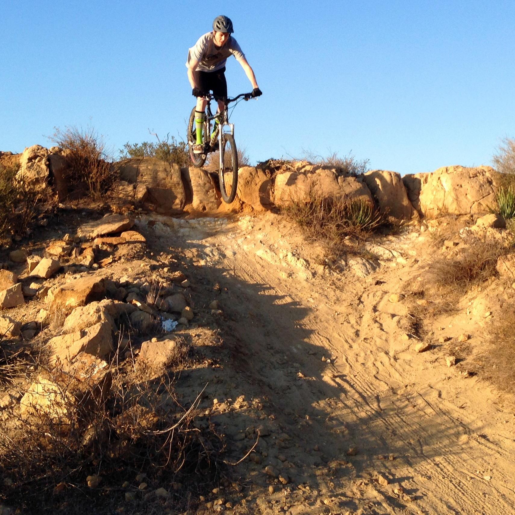 A person on a mountain bike is airborne while jumping off a rocky ledge, against a clear blue sky. The terrain is a mix of sand and rocky ground, with sparse vegetation in the background. Santiago Oaks mountain bike trail.