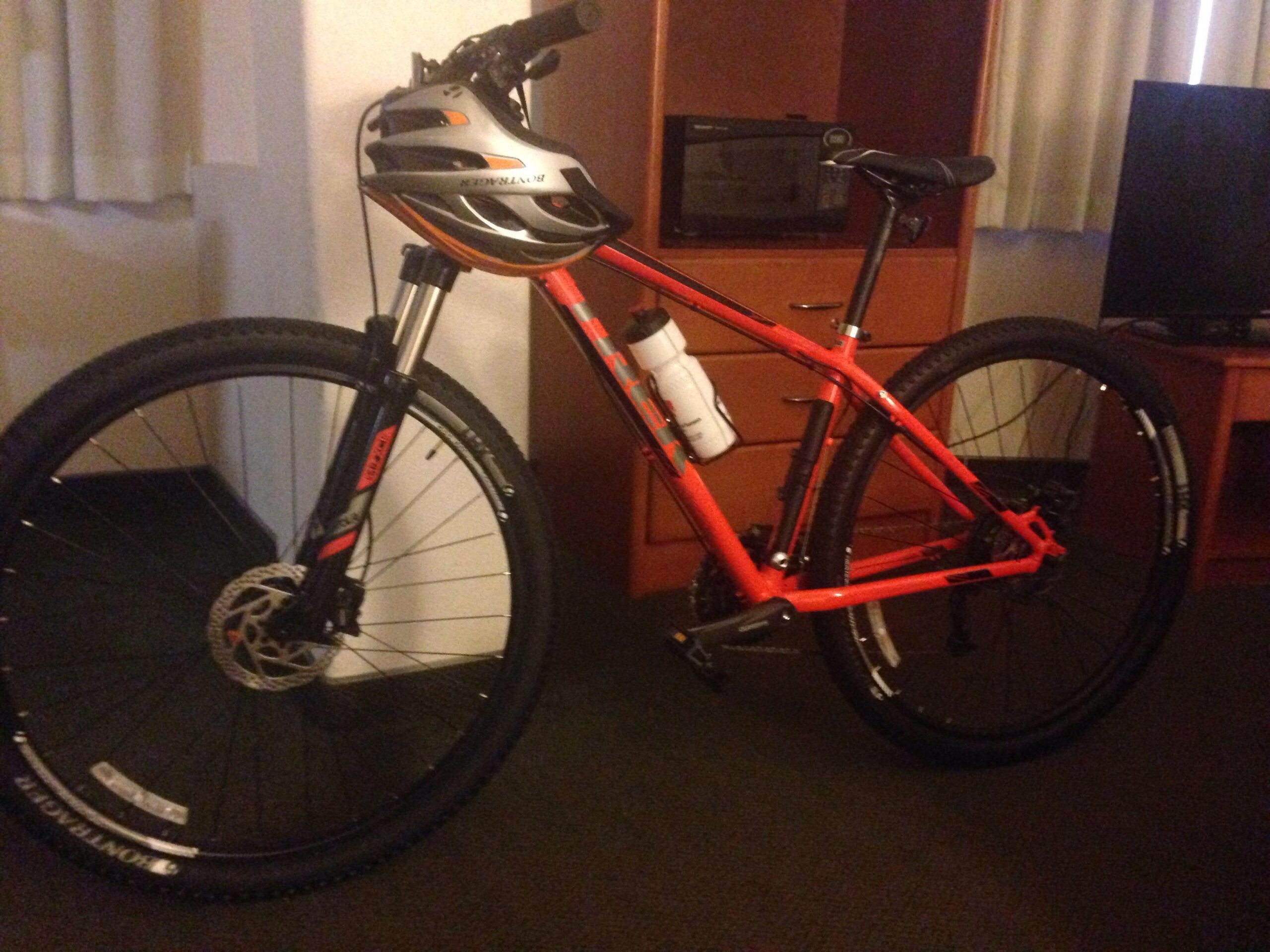 Trek X-Caliber 7: A vibrant orange mountain bike with thick tires and a water bottle mounted on the frame, positioned in a room that features wooden furniture and a television. A helmet rests on the handlebars, and there is a microwave visible in the background.