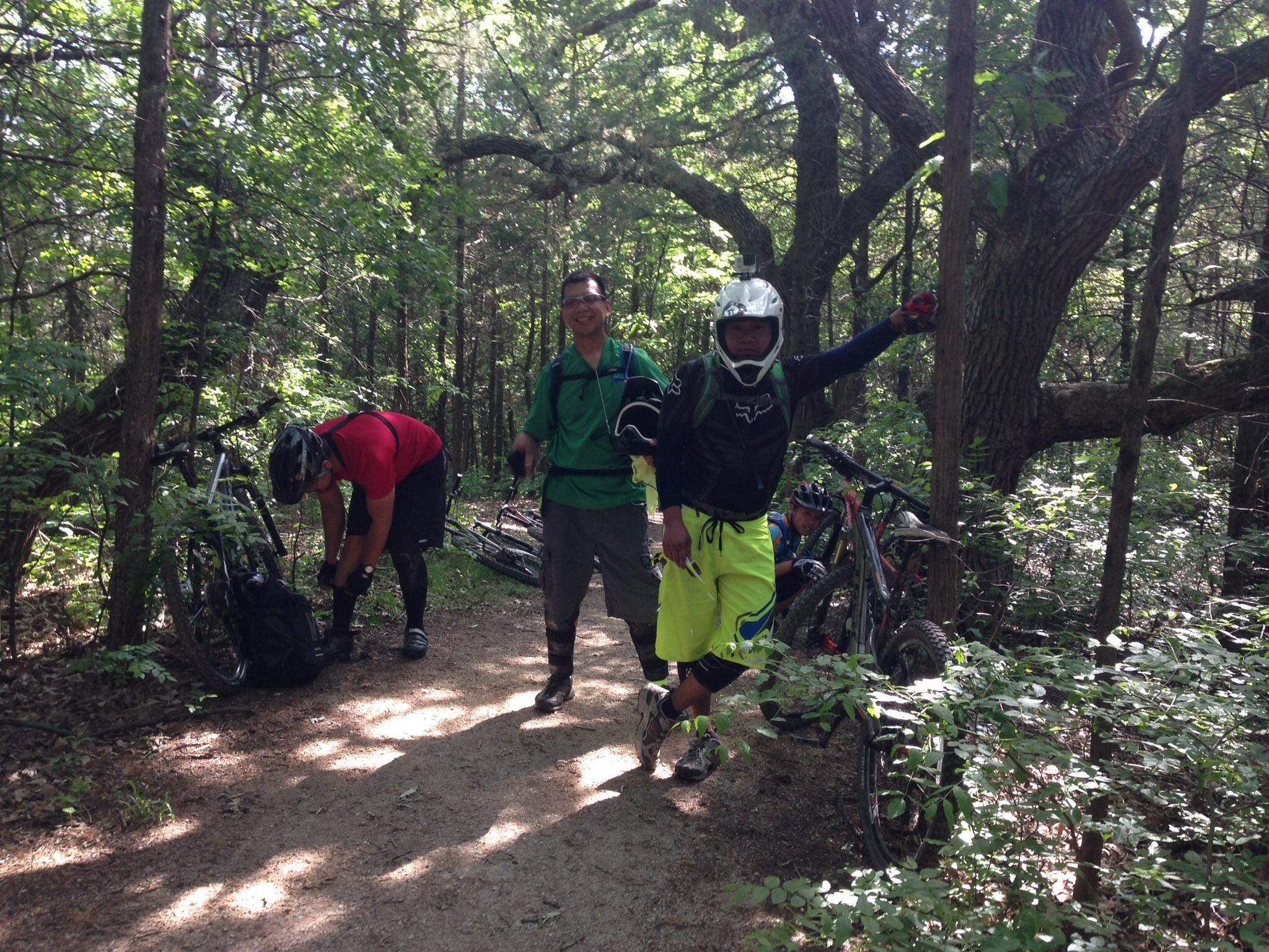 A group of three mountain bikers taking a break on a shaded dirt path in a forested area. One rider is leaning against a bike, smiling and gesturing, while another is adjusting gear in a backpack. The third is sitting nearby, also with a bike, surrounded by lush green foliage. Sunlight filters through the trees, creating a lively outdoor atmosphere. Emma Carlin mountain bike trail.