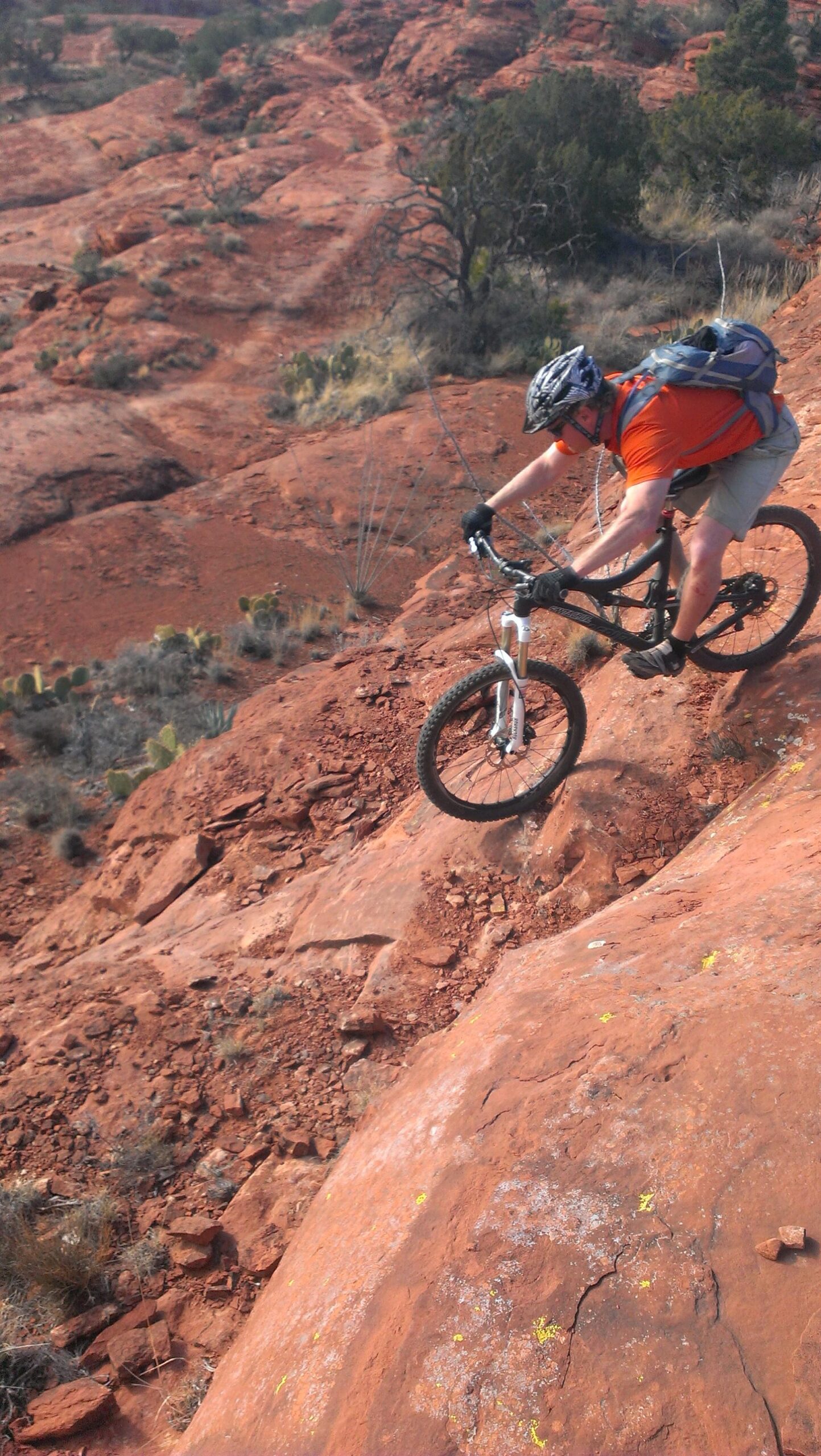 A mountain biker in an orange shirt navigates a rocky, red terrain, showcasing their skills on a steep downhill trail. The landscape features sparse vegetation and rugged rock formations. Hiline mountain bike trail.
