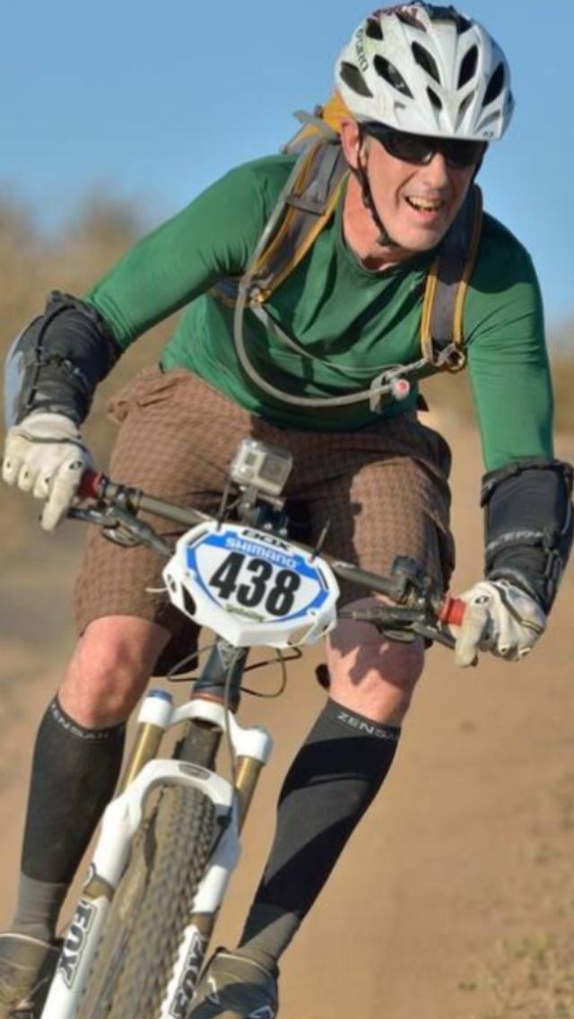 A mountain biker in a green long-sleeve shirt and brown shorts navigates a dirt trail, wearing a helmet and protective gear. The bike features a race number 438 on the front. The rider is smiling, conveying a sense of enjoyment and excitement while riding. Lake Hodges mountain bike trail.