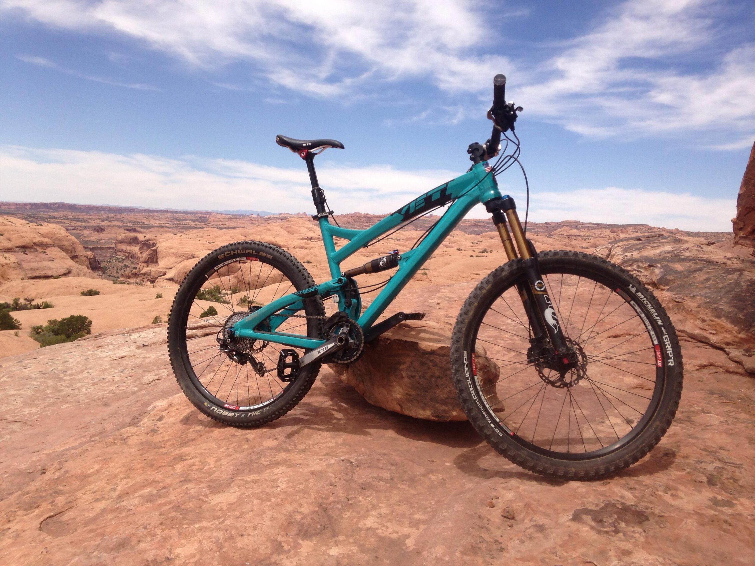 A turquoise mountain bike is leaning against a large rock in a rocky desert landscape, with a clear blue sky and distant hills visible in the background. Slickrock mountain bike trail.