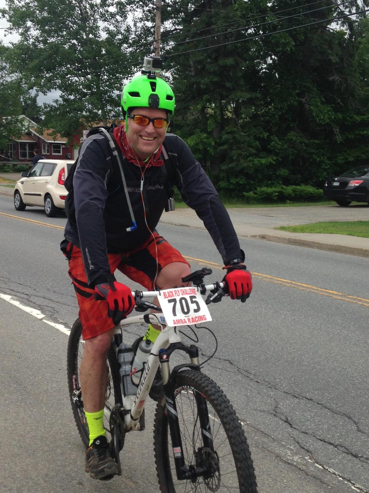 Specialized Stumpjumper Comp: A smiling cyclist wearing a bright green helmet, sunglasses, and red gloves rides a mountain bike on a road. He is dressed in a black long-sleeve shirt and red plaid shorts, and has a race number (705) displayed on his bike. A camera is mounted on his helmet, and he has a hydration pack on his back. The background features trees and parked cars.