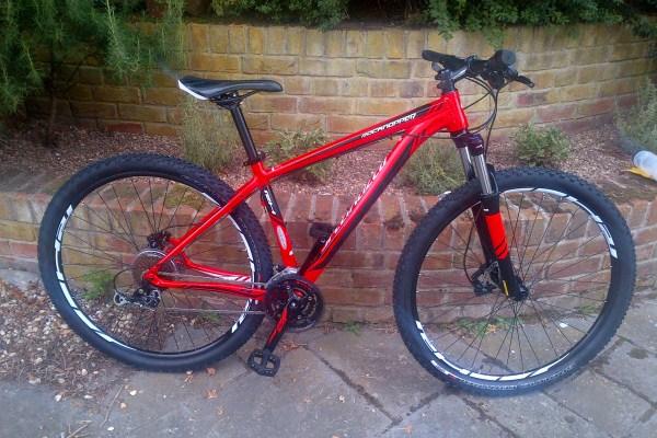 Specialized Rockhopper 29: A red mountain bike with wide tires and a suspension fork is leaning against a brick wall surrounded by greenery. The bike features a sleek design with a prominent frame and gear components.
