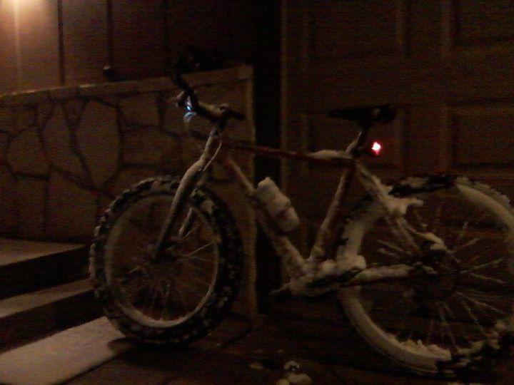 Surly Karate Monkey: A bicycle partially covered in snow, leaning against a porch at night. The bike features thick tires designed for winter riding and has a small light attached to the handlebars. The background includes a stone wall and a garage door.