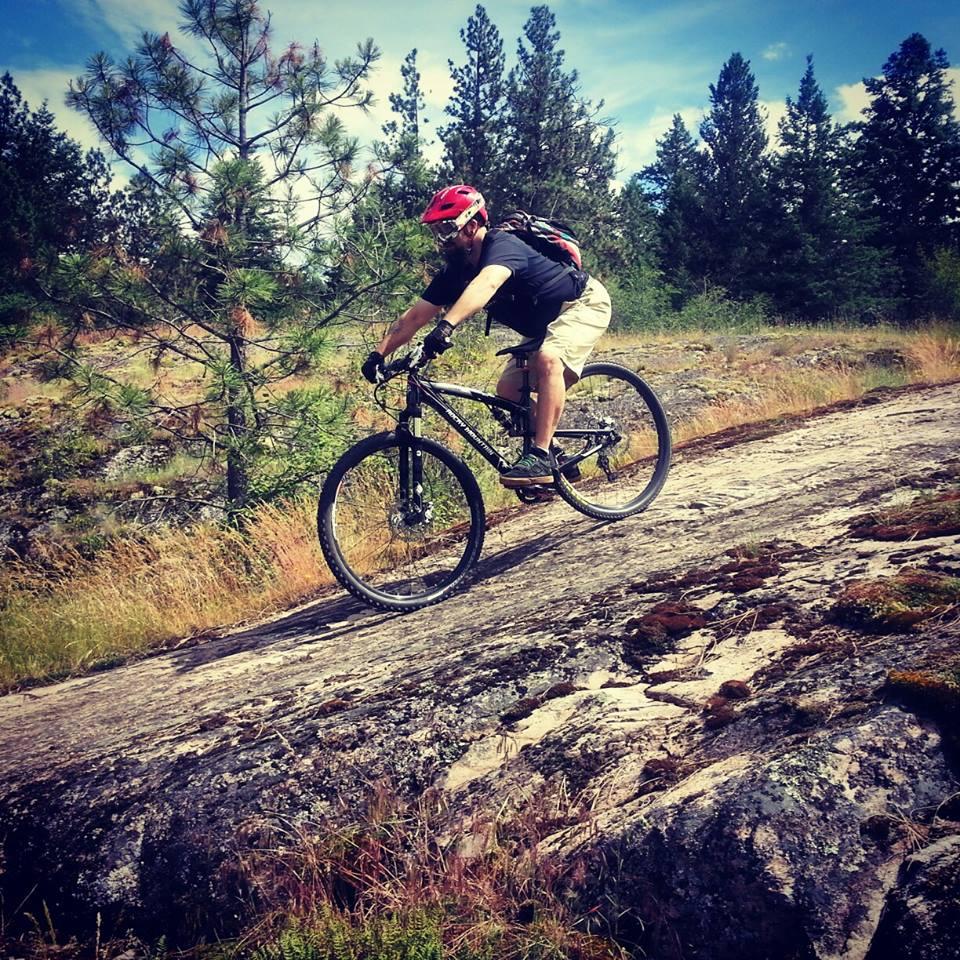 A person riding a mountain bike on a rocky trail surrounded by grass and pine trees on a sunny day. The cyclist is wearing a red helmet, black t-shirt, and beige shorts, navigating a sloped rock surface. Kalamalka Lake Provincial Park mountain bike trail.