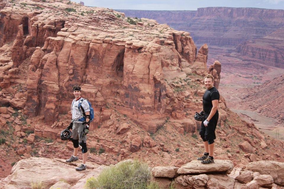 Two mountain bikers stand on rocky outcrops overlooking a dramatic canyon landscape. The reddish rock formations rise steeply in the background, while the foreground features shrubs and smaller rocks. One biker is dressed in a patterned jersey and protective gear, holding a helmet, while the other is in a black shirt and shorts, looking toward the camera. The sky above is partly cloudy, adding to the adventurous atmosphere of the scene. Amasa Back Trail mountain bike trail.