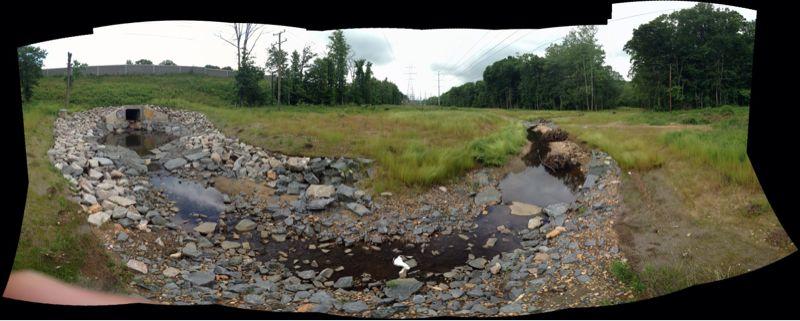 A panoramic view of a natural landscape featuring a shallow stream bordered by rocky banks. The scene includes patches of grass and sparse trees in the background, with a cloudy sky above. Power lines can be seen in the distance, indicating a human influence on the rural setting. Wakefield mountain bike trail.