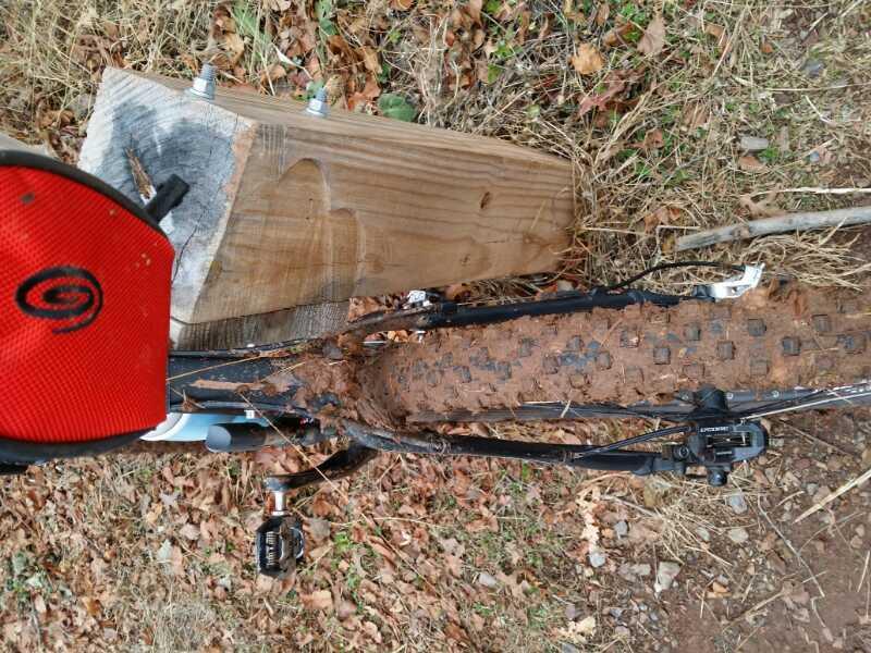 A close-up image of a mountain bike partially covered in mud, resting against a wooden post. The bike's rear wheel features a textured tire, and a red cap is visible in the foreground, suggesting recent activity in a natural, outdoor setting with dry leaves on the ground. Six Mile Run mountain bike trail.