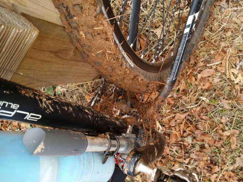 A close-up view of a mountain bike with a muddy rear tire, showcasing mud caked on the wheel, chain, and frame. The ground is covered with fallen leaves, and a portion of a wooden structure is visible in the background. Six Mile Run mountain bike trail.