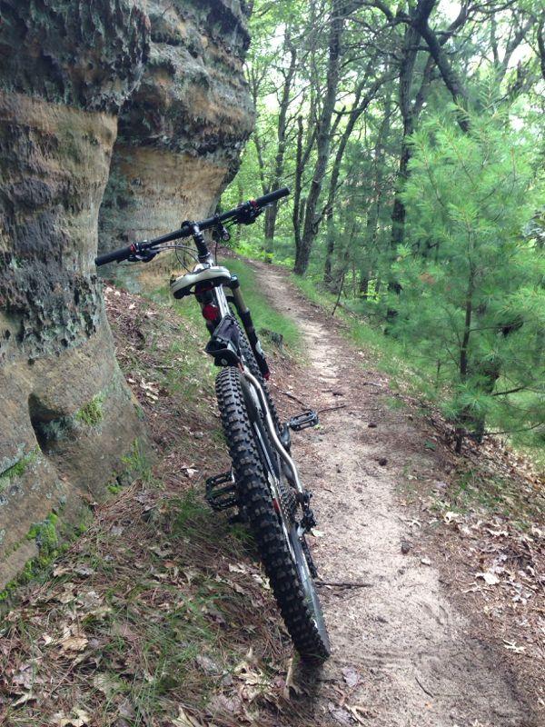 A mountain bike leaning against a rocky cliff on a dirt path surrounded by lush greenery and trees. Levis Mounds mountain bike trail.
