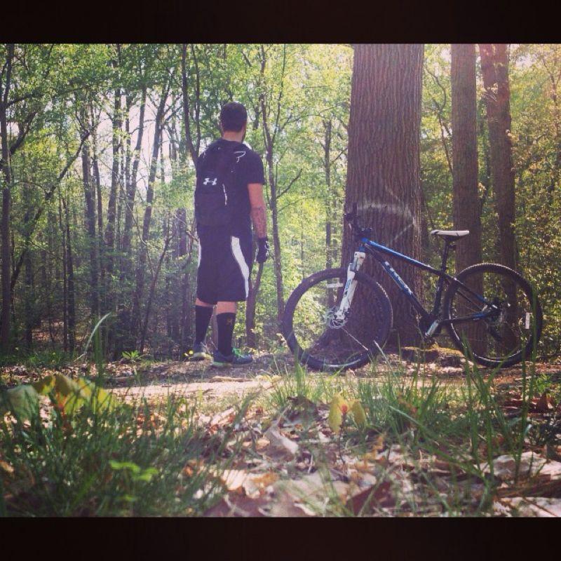 A person standing in a wooded area, facing away from the camera, wearing a black shirt, shorts, and a backpack. Next to them is a blue mountain bike resting against a tree. The setting is lush with greenery, showcasing trees and foliage in a sunny environment. Camden County College mountain bike trail.