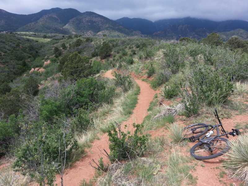 A winding dirt trail through a lush landscape with scrubs and grasses, leading into the mountains under a cloudy sky. A mountain bike is lying on its side nearby, suggesting recent use along the trail. Red Rock Canyon mountain bike trail.
