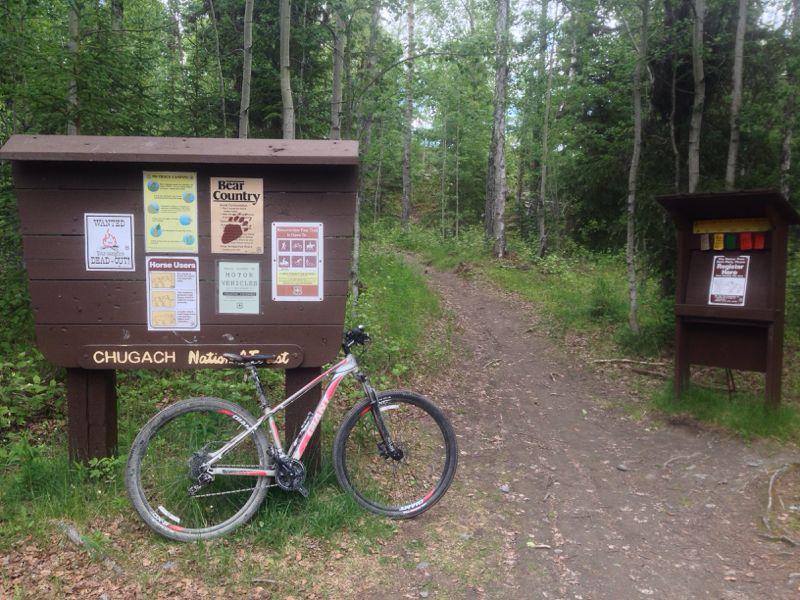 A mountain bike leans against a wooden signboard at the entrance to a trail in Chugach National Forest. The sign features various informational posters, including warnings about wildlife, trail regulations, and activity guidelines. The lush, green forest surrounds the trail, leading into the wilderness. Another smaller information board stands on the right. Resurrection Pass mountain bike trail.