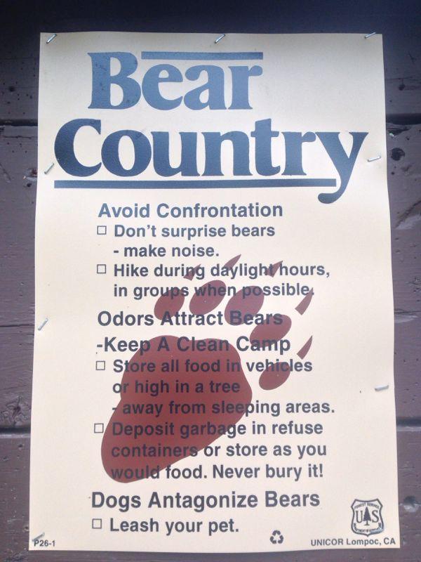 A warning sign titled "Bear Country," providing safety tips for outdoor activities in areas inhabited by bears. The list includes recommendations to avoid bear encounters, such as making noise to avoid surprising bears, hiking during daylight, and storing food properly to minimize odors. It also advises keeping a clean campsite and leashing pets to prevent attracting bears. The sign features a bear paw print graphic and is printed on yellow paper. Resurrection Pass mountain bike trail.