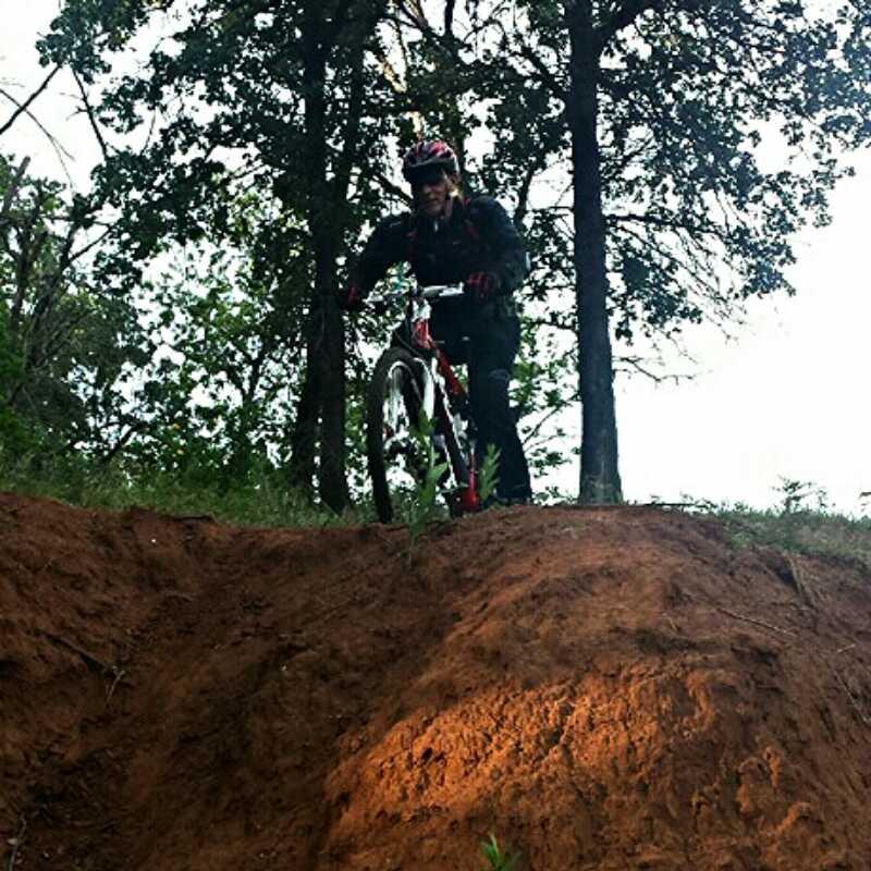 A mountain biker stands on a dirt trail with a steep slope in front of them, surrounded by trees. The cyclist is wearing a helmet and riding gear and is poised to descend the incline on a red and white mountain bike. The scene is set in a natural outdoor environment. Eagle View, Kaw Lake mountain bike trail.