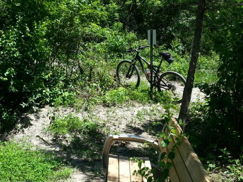 A black mountain bike leaning against a post in a lush green area, with a wooden bench in the foreground. The scene includes dense foliage and a sandy path, creating a peaceful outdoor setting. Wildlife Prairie Park mountain bike trail.